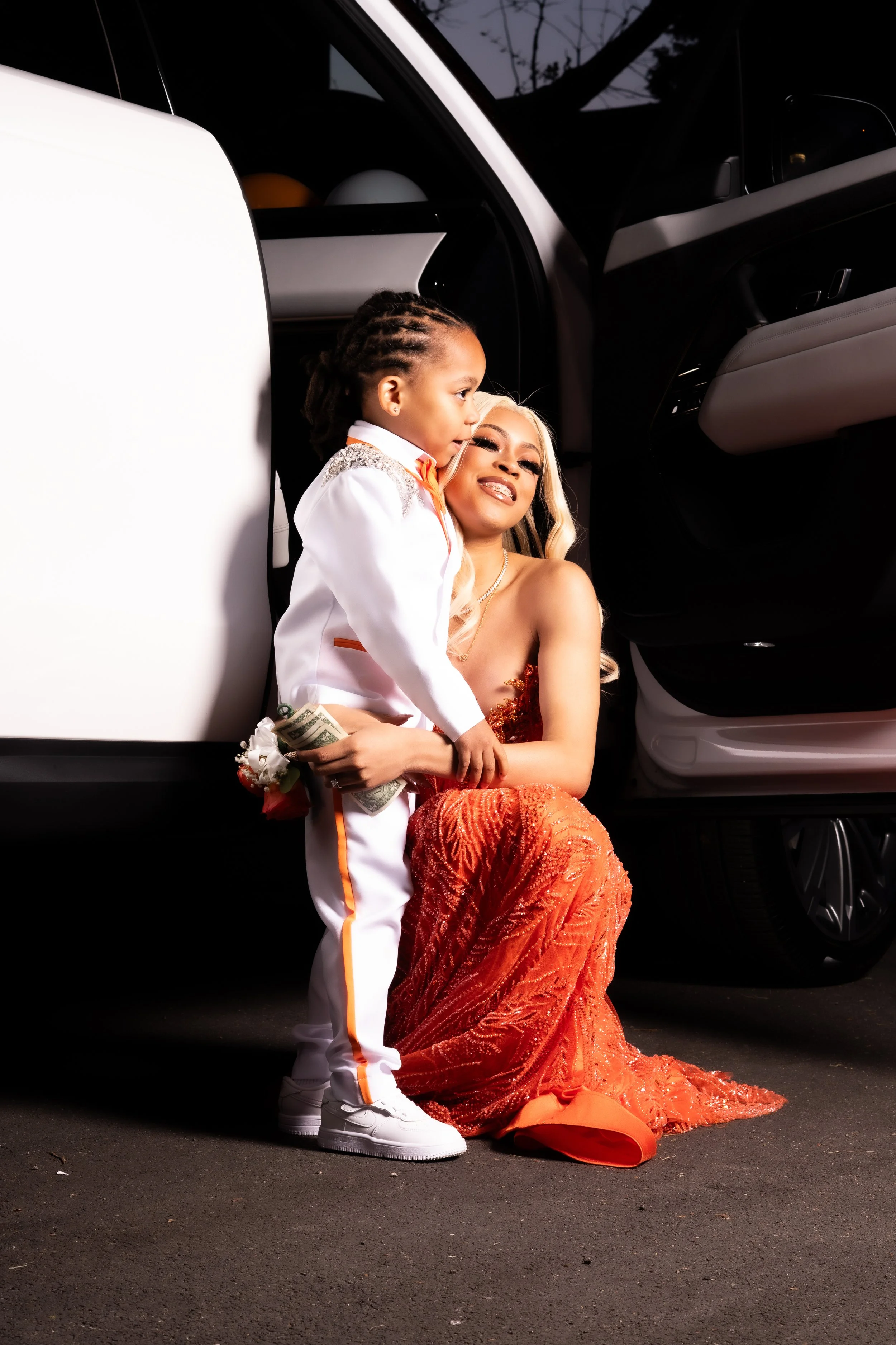 A woman in a glamorous orange dress and a young boy in a white tuxedo with orange accents are posing together in front of a black car at night. The boy is holding dollar bills, and the woman is sitting on the ground with a smile, while the boy is sta