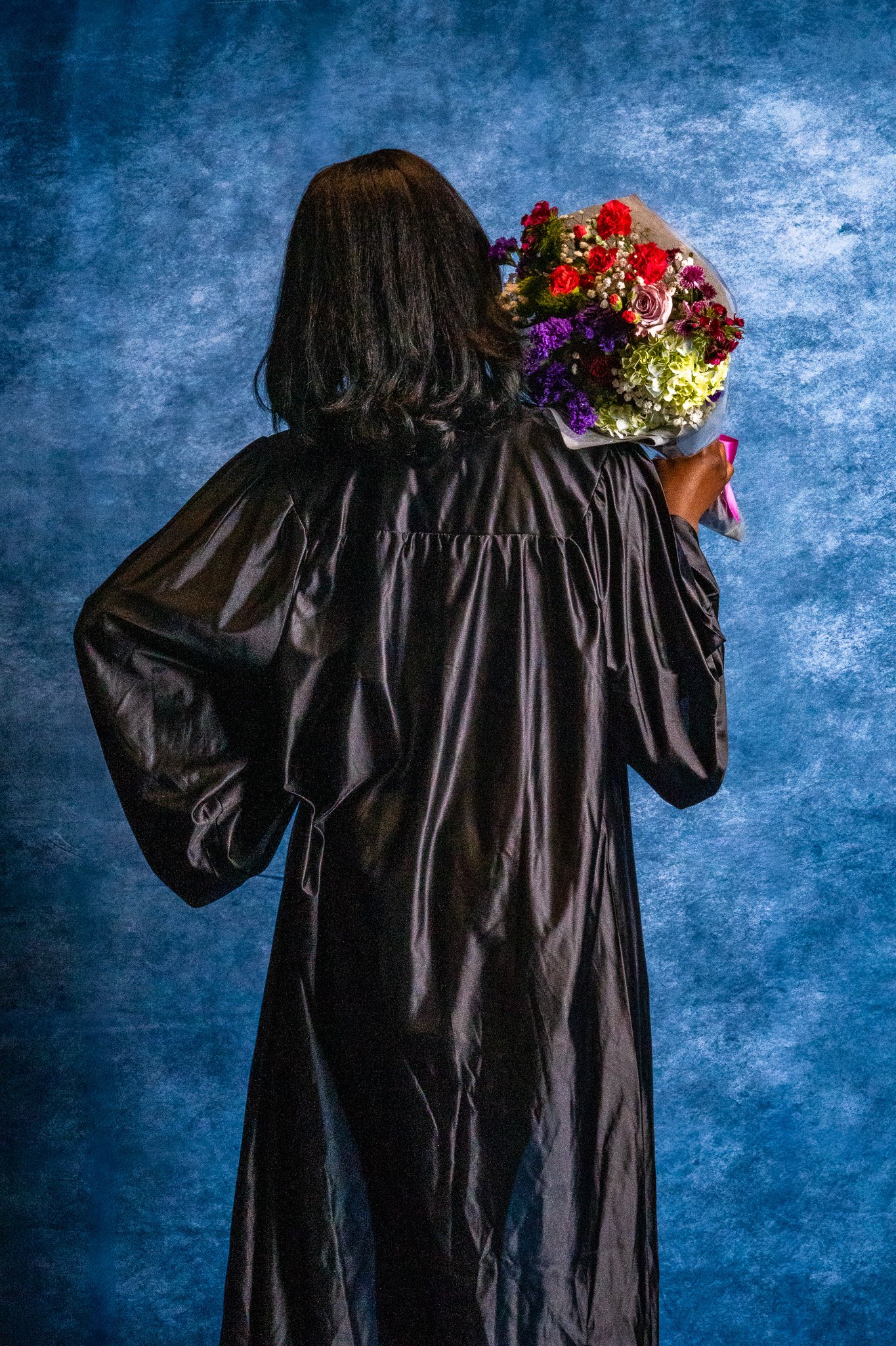 A person with black hair, wearing a black graduation gown, is facing away and holding a large bouquet of colorful flowers against a blue textured background.