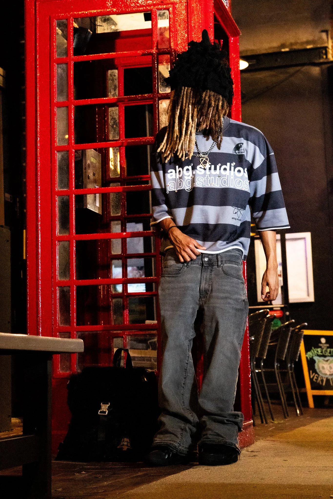 A person with dreadlocks wearing a black hat and striped shirt standing near a red telephone booth in a dimly lit indoor setting.