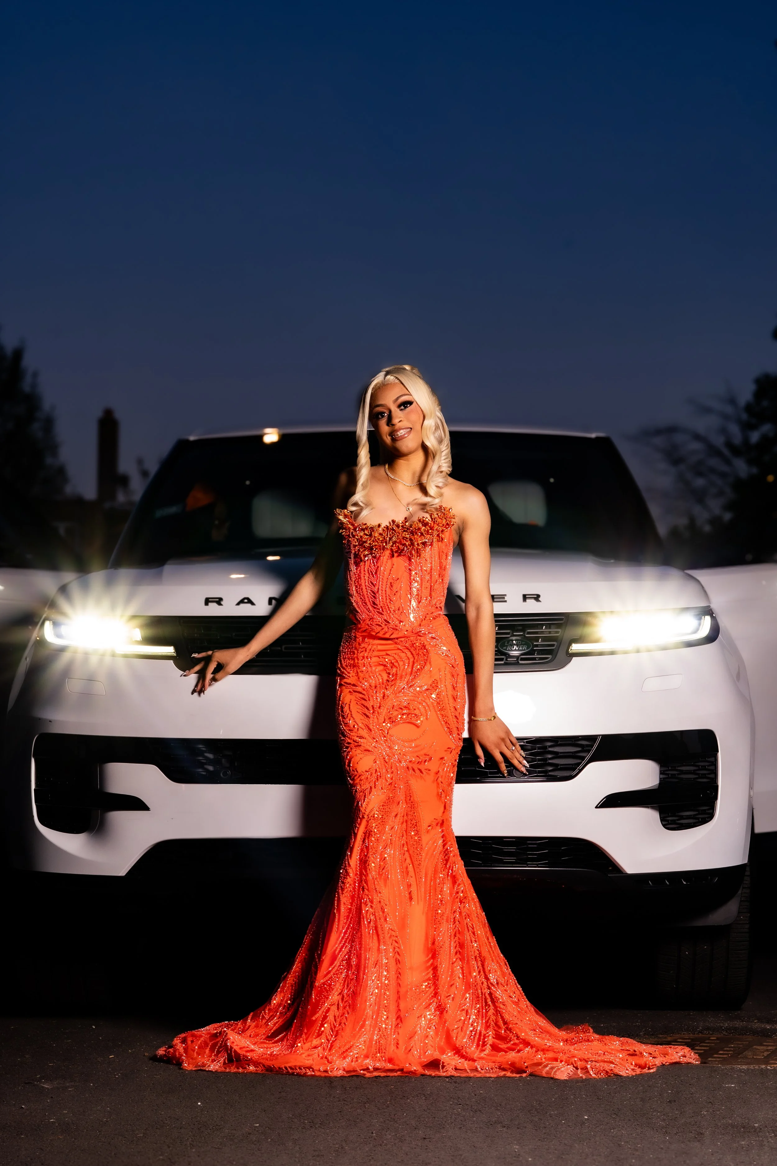 A woman in an elegant, fitted red dress with intricate beading and embroidery stands in front of a white Range Rover at night with a dark sky. The woman is smiling, has long blonde hair styled in loose curls, and is accessorized with necklaces and br