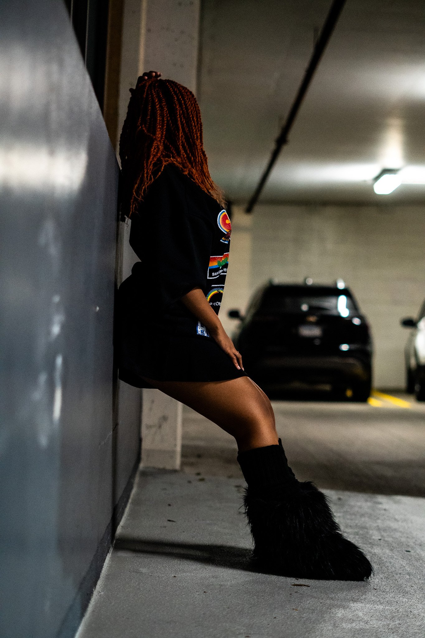 A person with red braided hair leaning against a wall in an indoor parking garage, wearing a black sweatshirt with patches, a black skirt, and black furry boots, with a black dog sitting nearby.