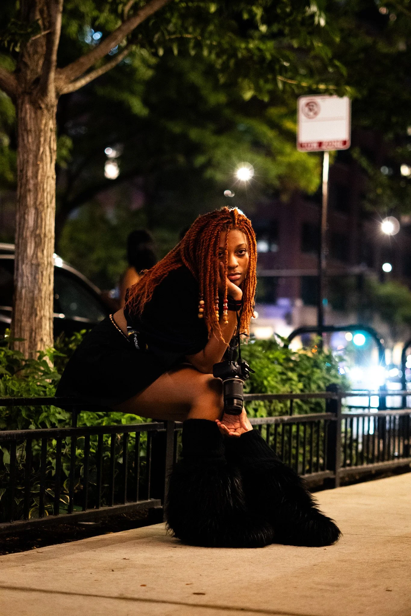 A young woman with long, orange dreadlocks sits on a park bench at night with city lights in the background. She is leaning on her hand, looking directly at the camera. A large black dog with shiny fur is sitting in front of her on the sidewalk.