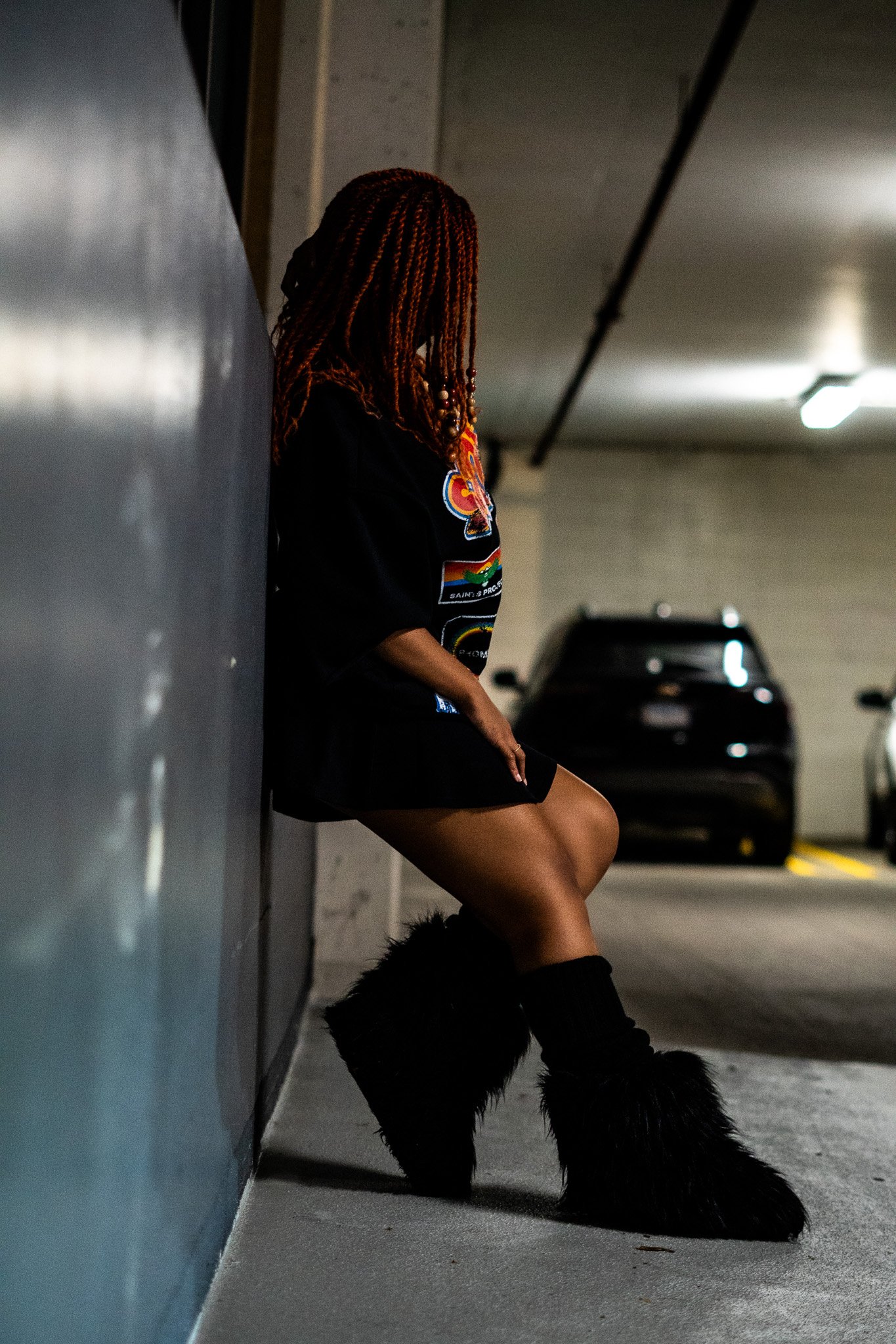 A person with red braids and a colorful shirt is leaning against a wall in a parking garage, sitting on the ground with legs crossed and furry black boots.