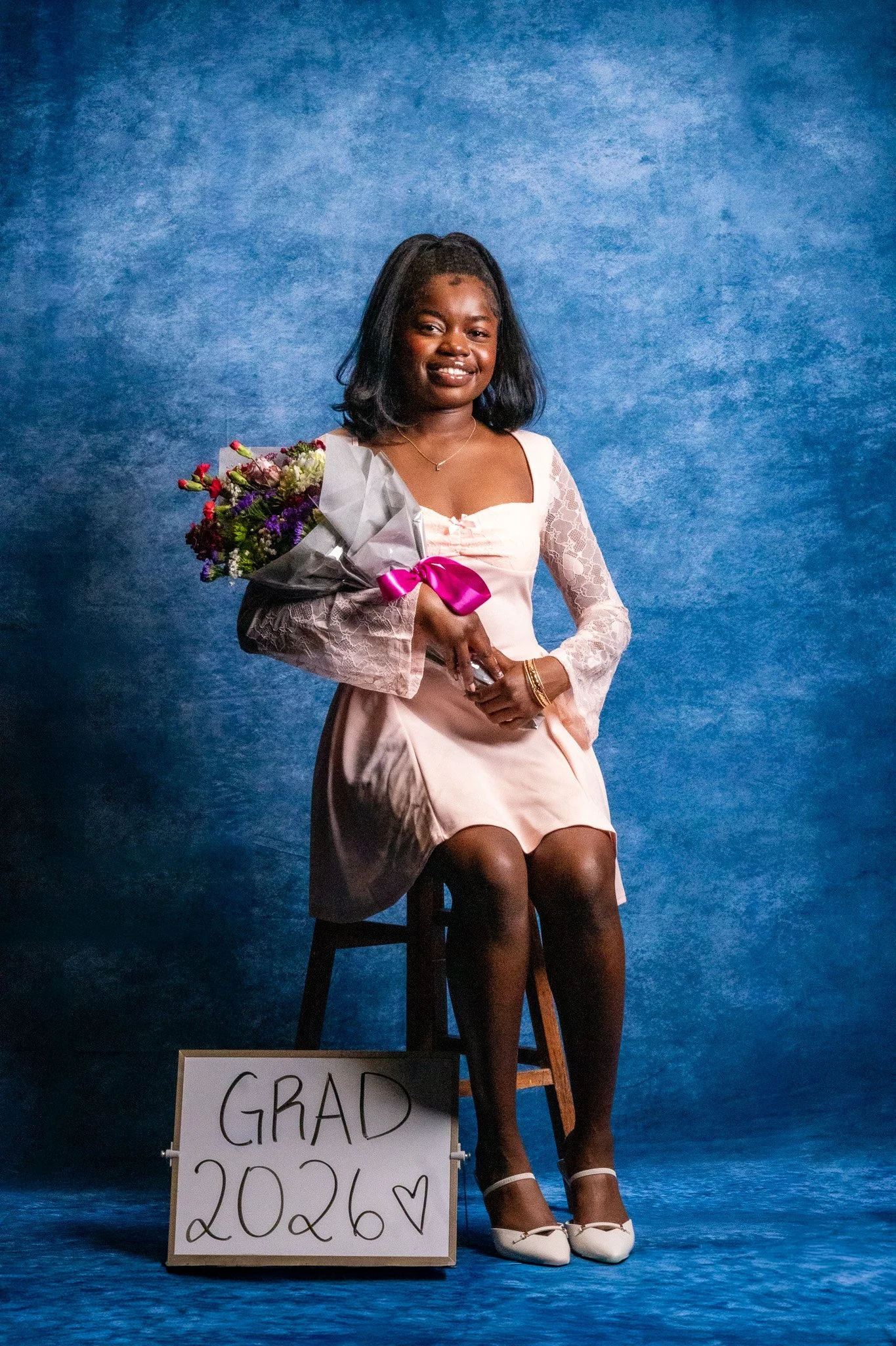 Young woman sitting on a stool, holding a bouquet of flowers, and wearing a pink dress with lace sleeves, white shoes, and gold jewelry, with a graduation sign on the floor that reads 'GRAD 2026' against a blue textured background.