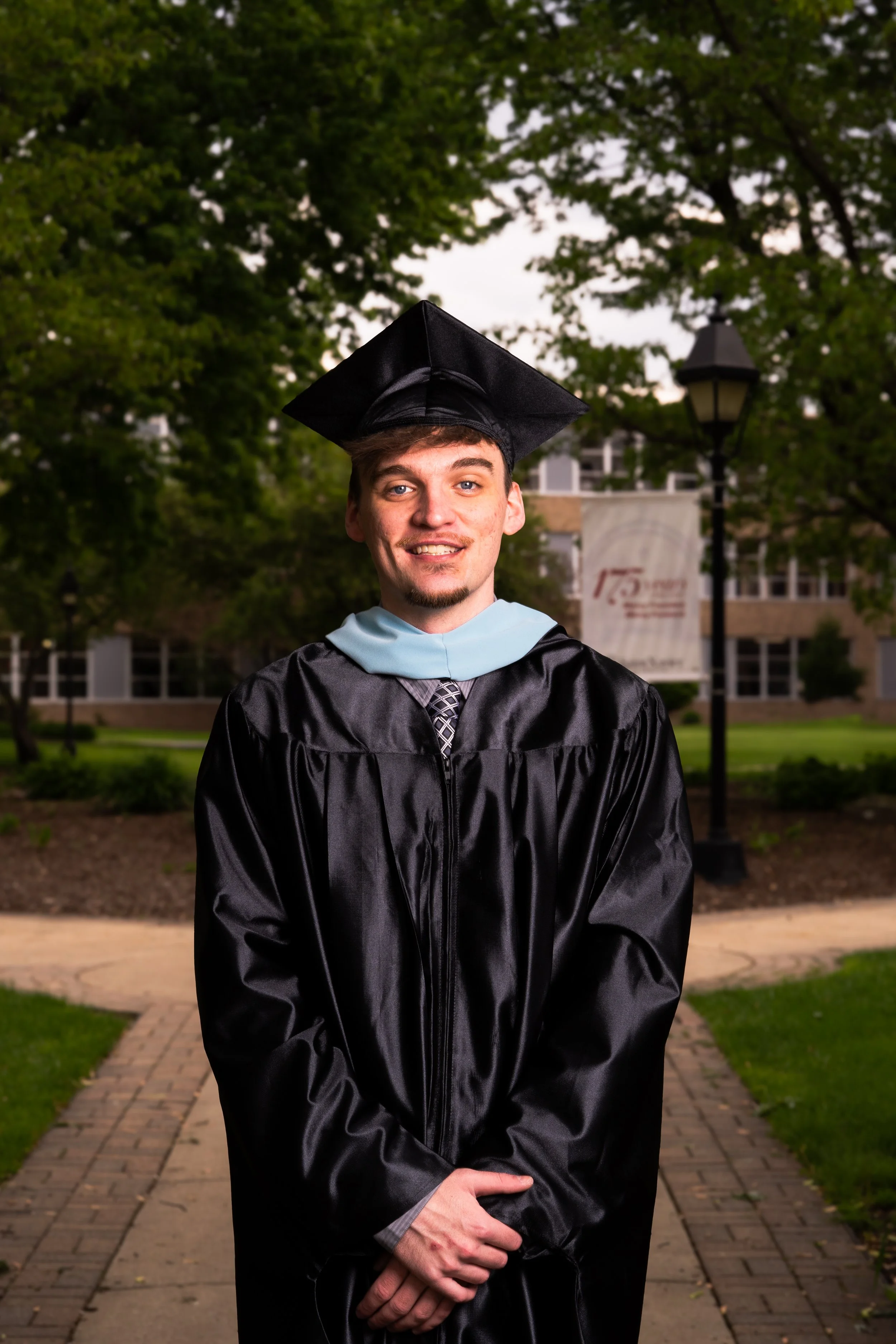 A young man wearing a black graduation gown and cap standing outdoors on a brick pathway with trees and buildings in the background, smiling.