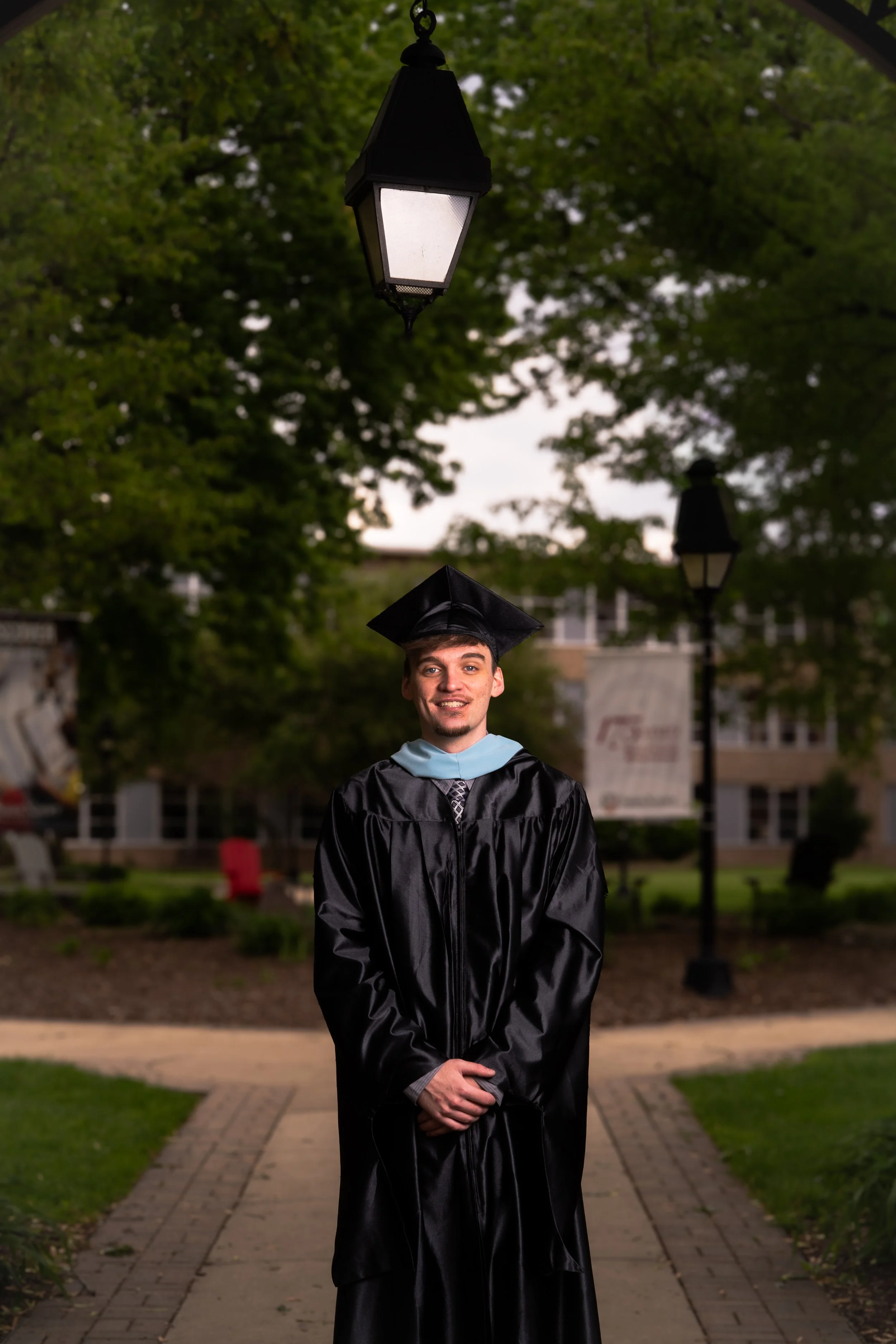 Young man in graduation cap and gown smiling outdoors in a park with trees and street lamps.