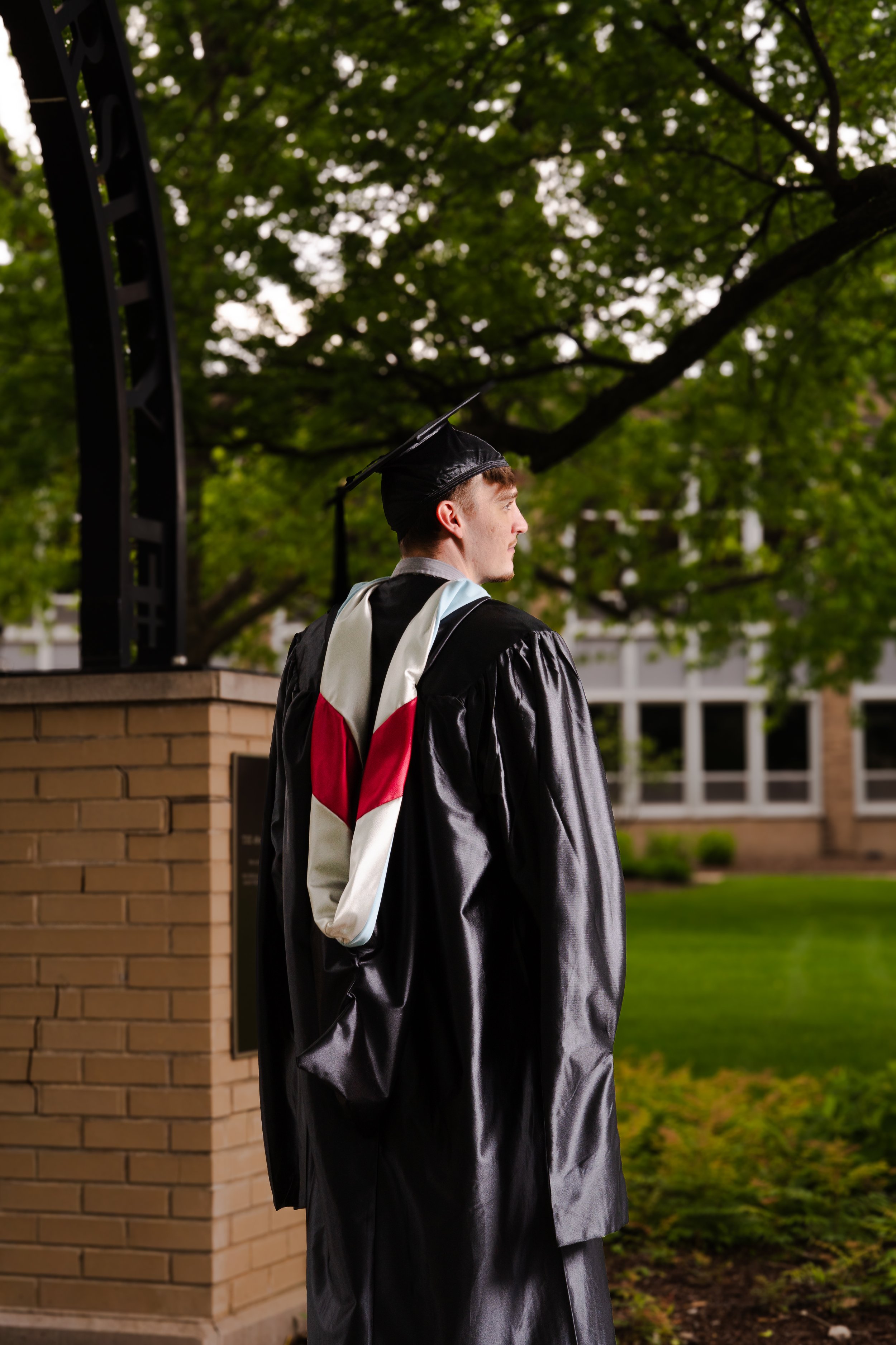 A young man in a black graduation gown and cap stands outdoors, looking to the right. He is wearing a white and red stole, with green trees and a brick building in the background.