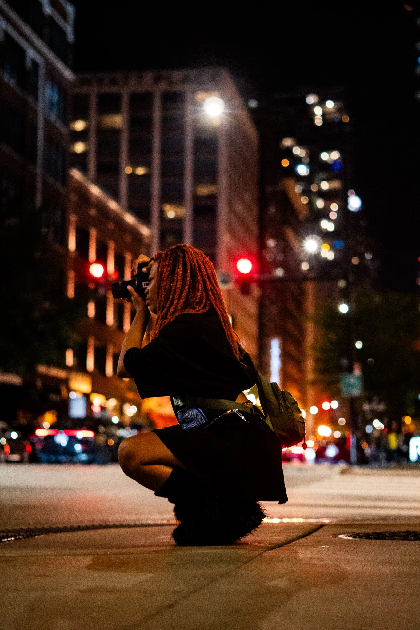 A woman with red braids kneeling at a crosswalk at night, holding a camera, with city buildings and busy street lights in the background.