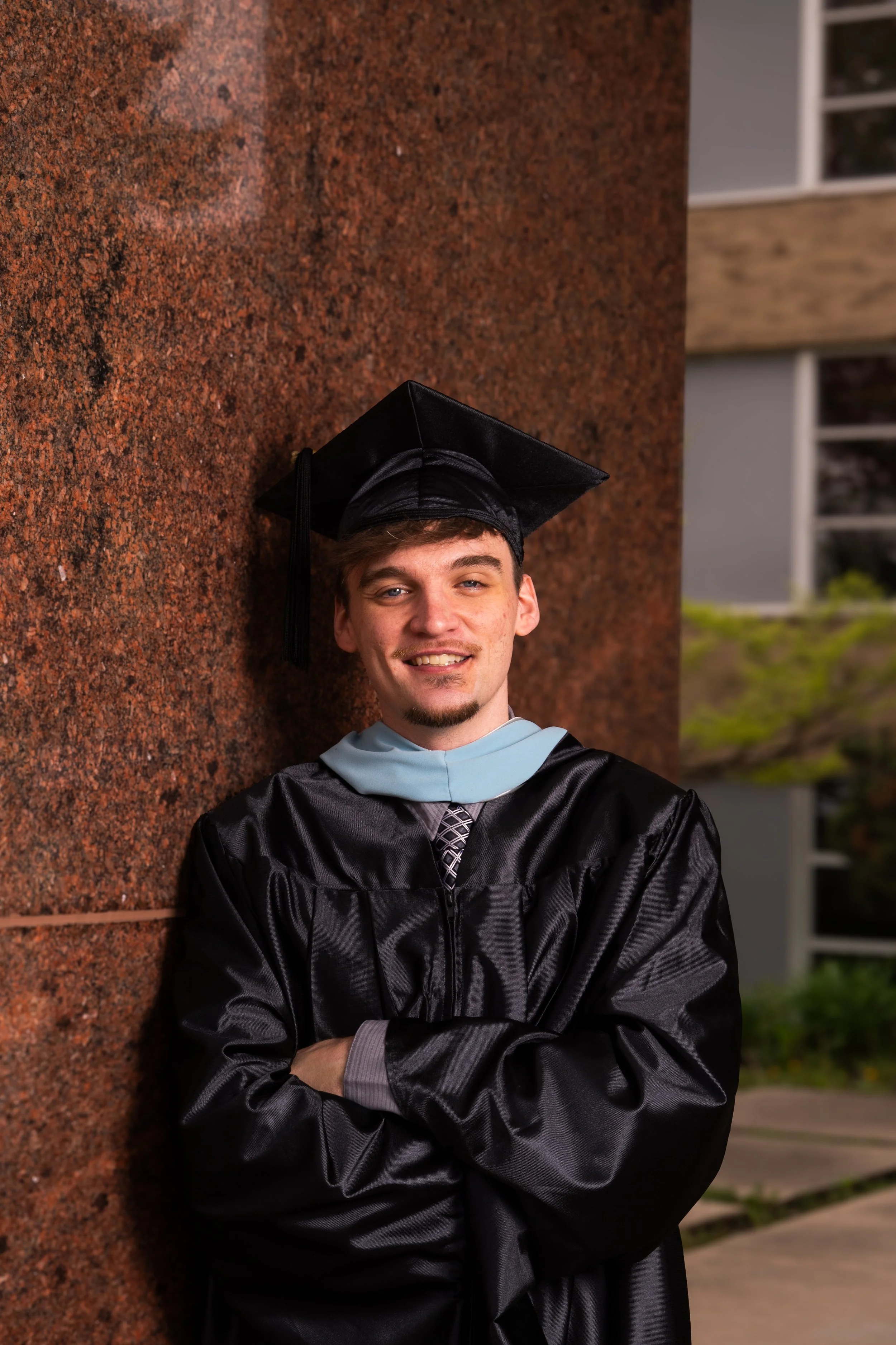 Young man in graduation cap and gown leaning against a brick wall outdoors, smiling.