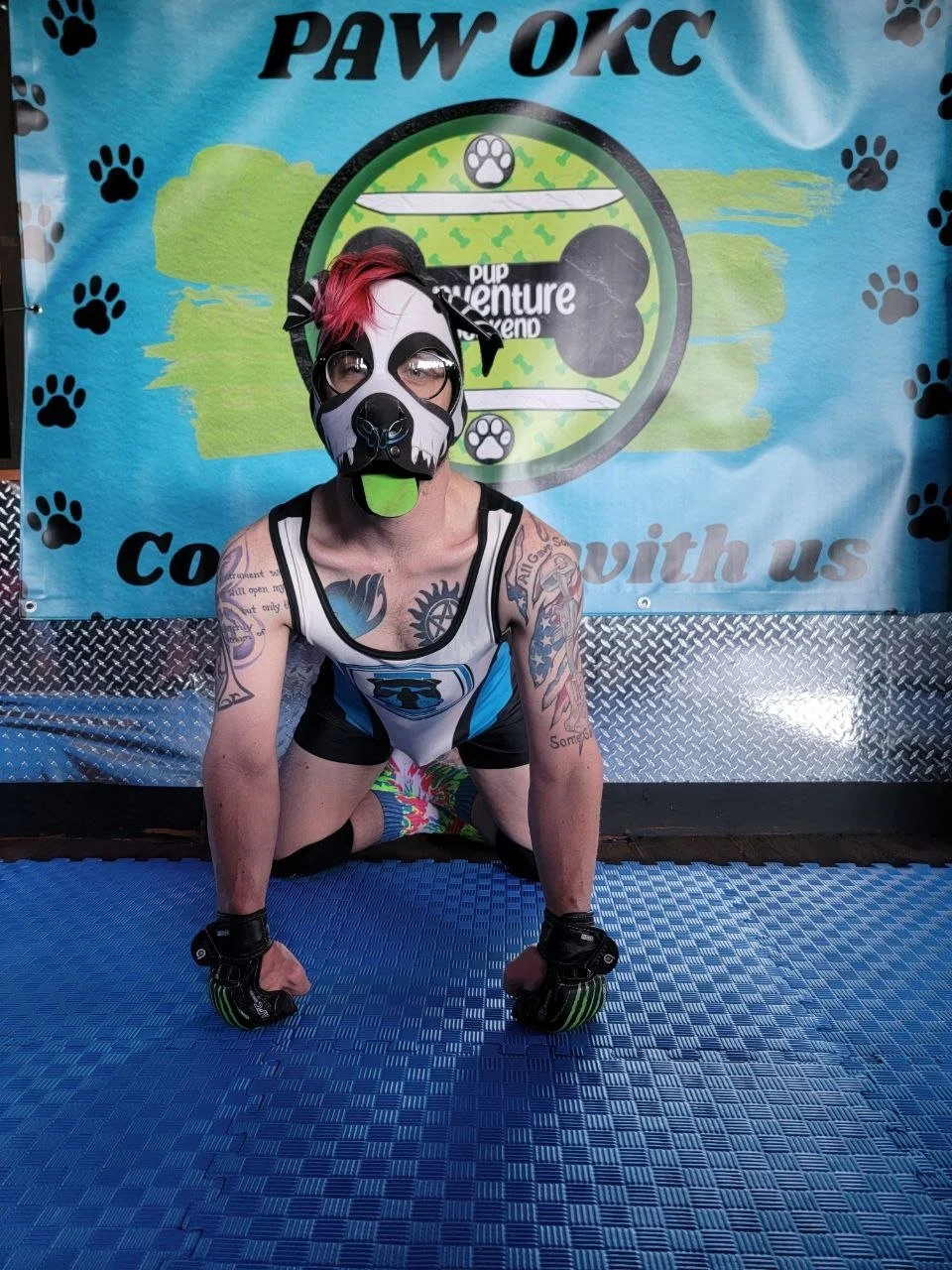 Person wearing a dog mask and workout gloves, crouching on a blue exercise mat in front of a colorful backdrop with paw prints and a dog paw logo, at a puppy adventure event.