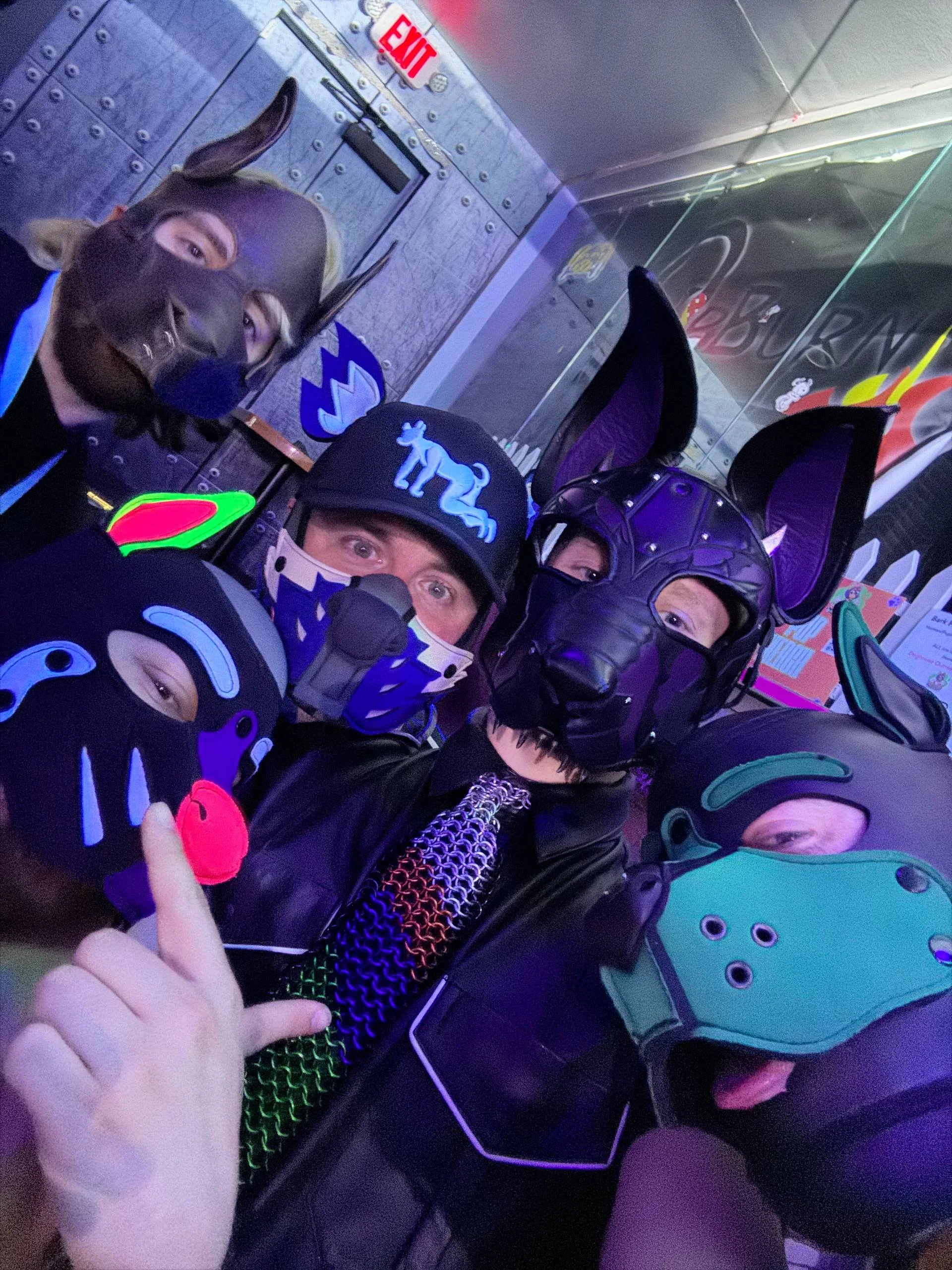 Three people wearing costumes with purple and black cat-like masks, some with horns or feline ears, in a dimly lit setting with colorful neon lights and gaming or entertainment posters in the background.