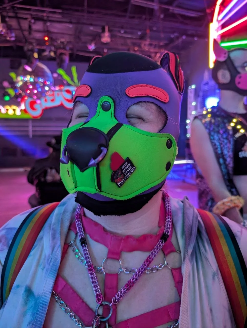 Person wearing a colorful, unicorn-themed mask with a green face covering and rainbow accents, in a neon-lit environment with other people.