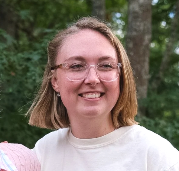 Smiling woman wearing clear glasses and a white shirt outdoors in a wooded area