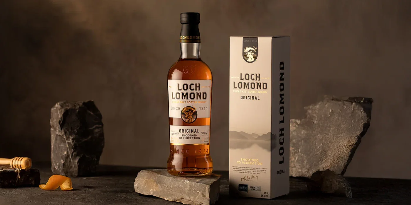 Botella y caja de whisky Loch Lomond en un fondo oscuro con rocas decorativas.