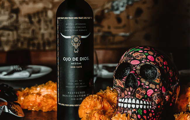 Botella de mezcal Ojo de Dios, decorada con cráneos de calavera y flores coloridas en un fondo rústico.