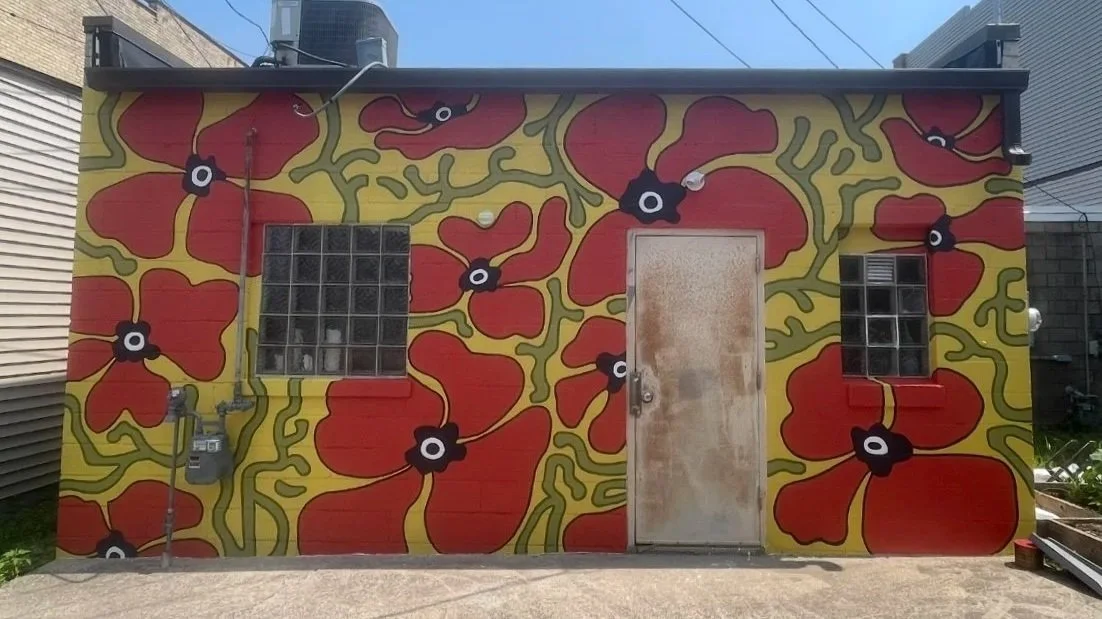 A building with exterior walls painted with a large, colorful floral mural featuring red flowers with black and white centers and yellow-green background accents.