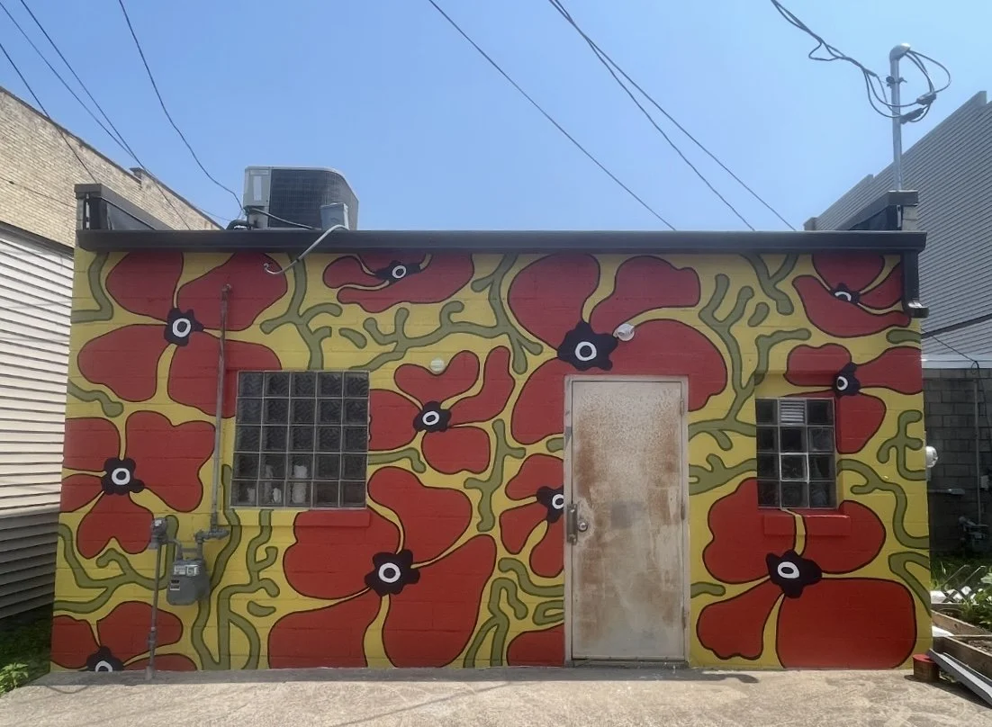 Building exterior painted with a red and yellow floral mural, featuring large red flowers with black and white centers, a small window made of glass blocks, a metal door, and utility wiring on the wall.