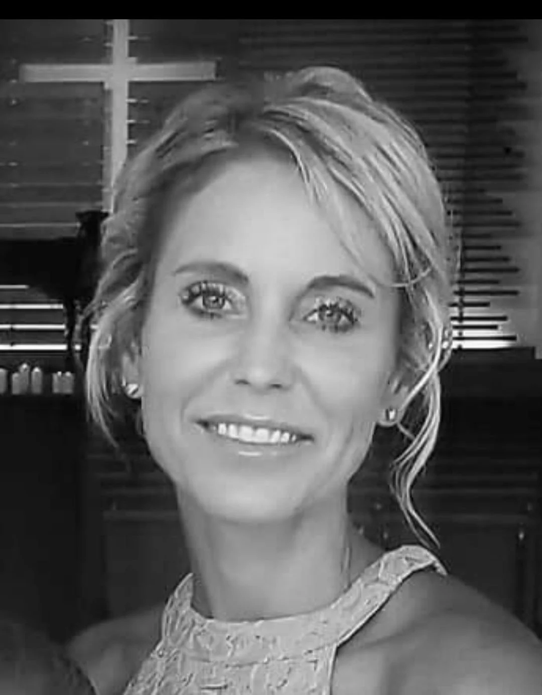 Black and white photo of a smiling woman with short, wavy hair. She is wearing earrings and a lace top. The background includes window blinds and a piano.