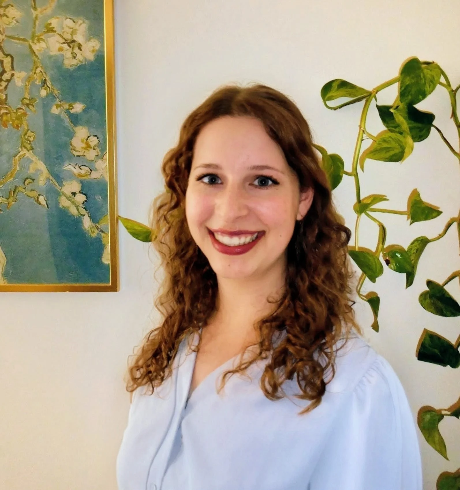 A young woman with long, curly red hair and blue eyes smiling, wearing a white top, standing in front of a wall with a map and a green plant with large leaves.