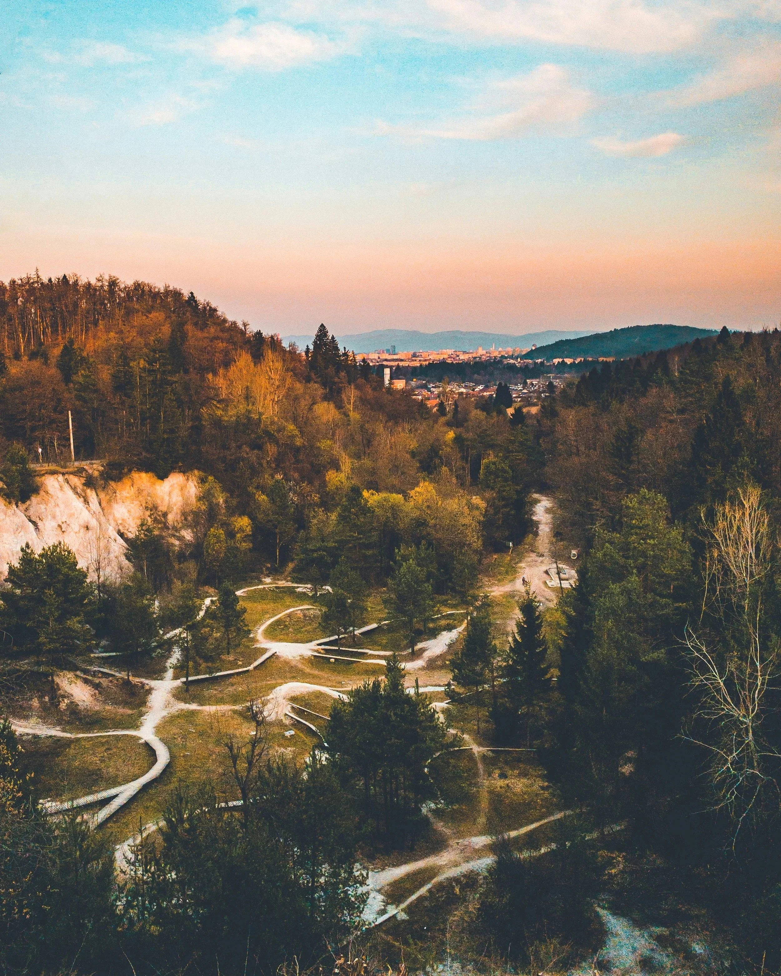 Hilly landscape with a winding road, trees, and a cityscape in the distance under a partly cloudy sky during sunset.