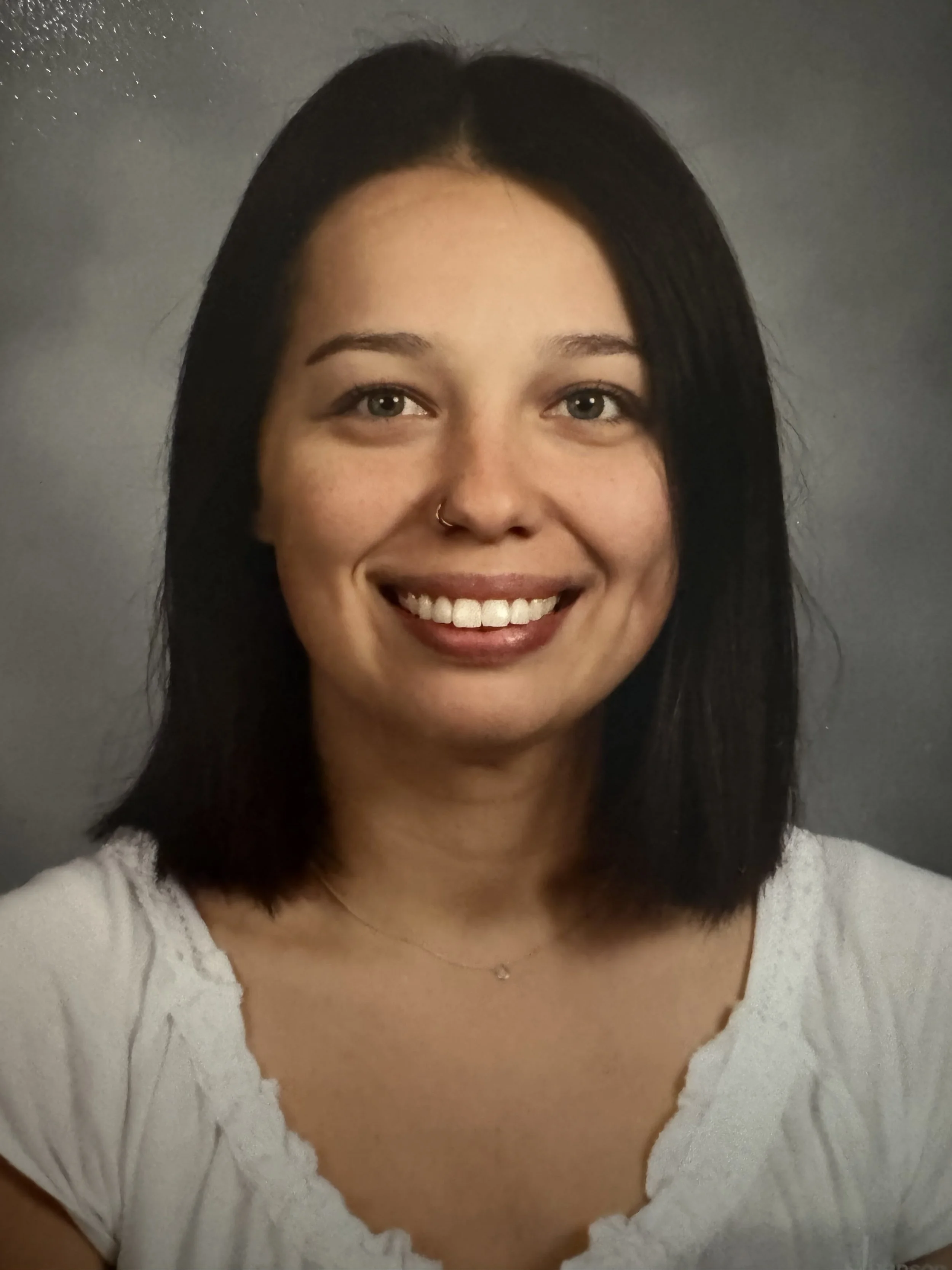 Close-up of a smiling woman with black hair, light skin, a nose ring, wearing a white top with lace details and a delicate necklace, against a dark background.