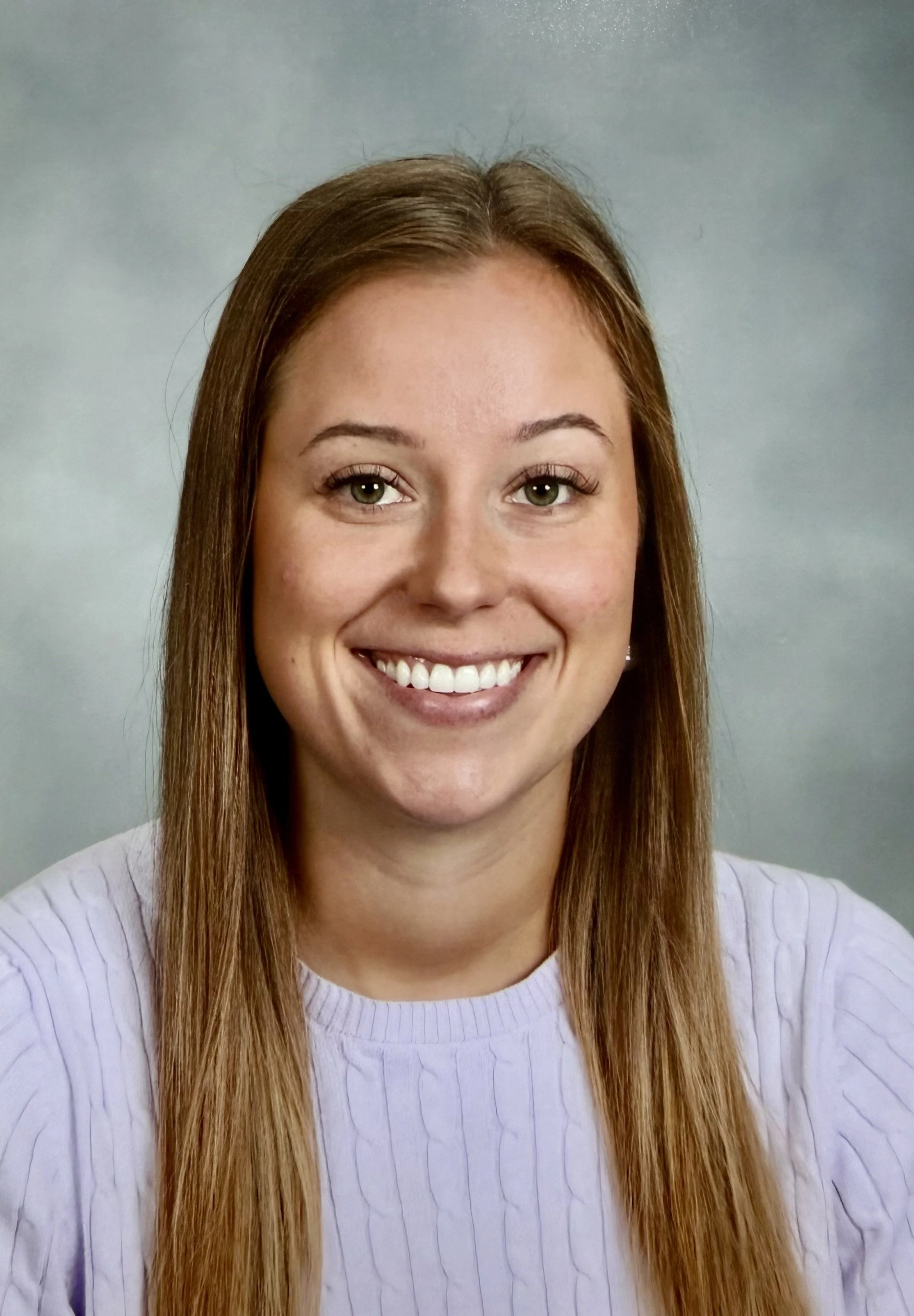 A woman with long, straight light brown hair smiling, wearing a light purple ribbed sweater, against a plain gray background.