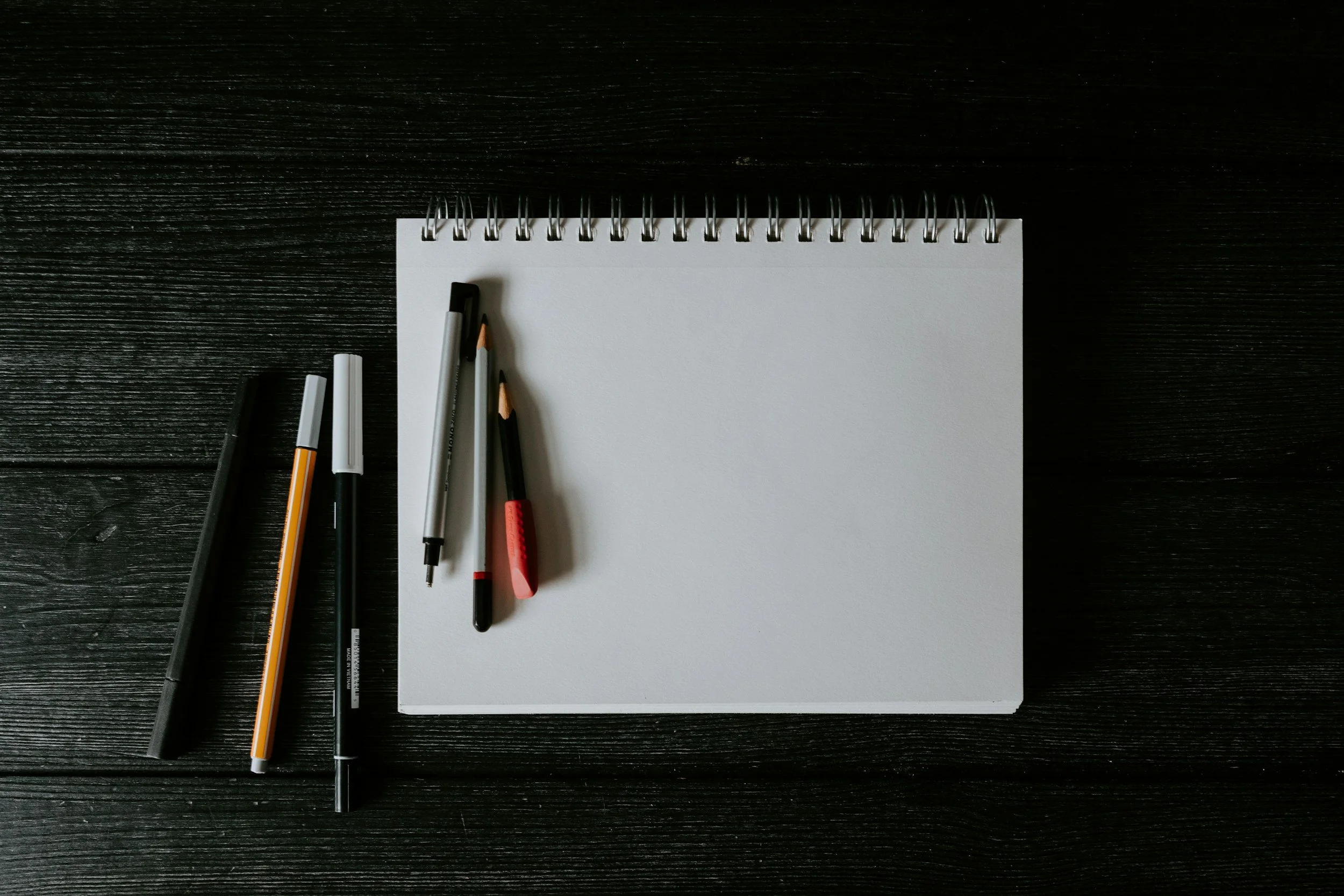 A blank spiral-bound notepad with several pens and markers placed on a dark wooden surface.