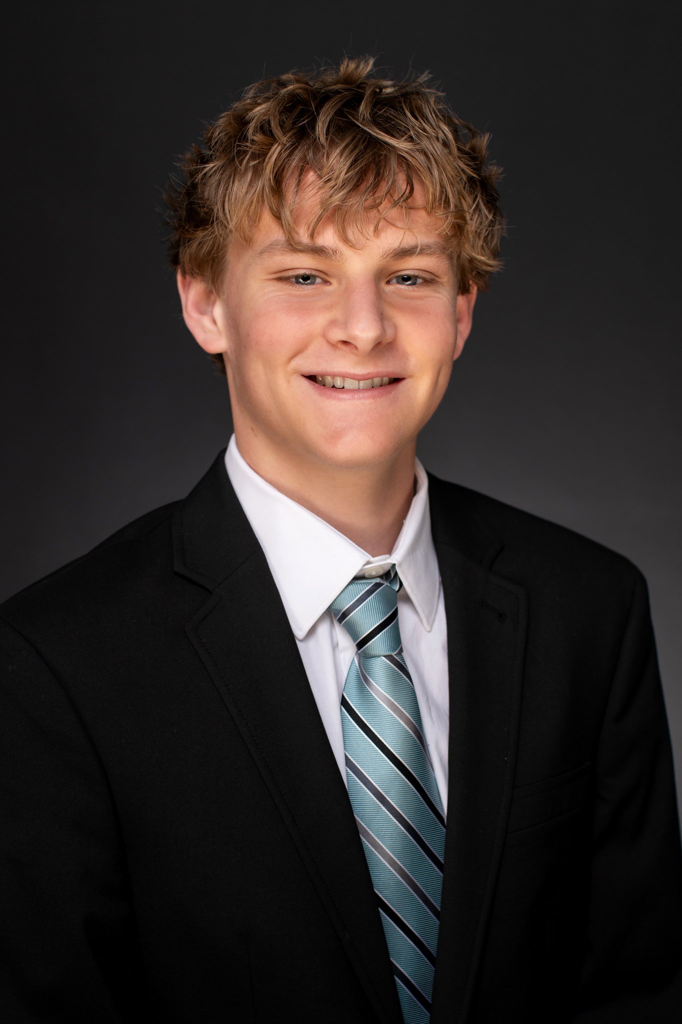 A young man with blonde, curly hair smiles and poses in formal attire, wearing a black suit jacket, white dress shirt, and a striped blue and white tie, against a dark background.
