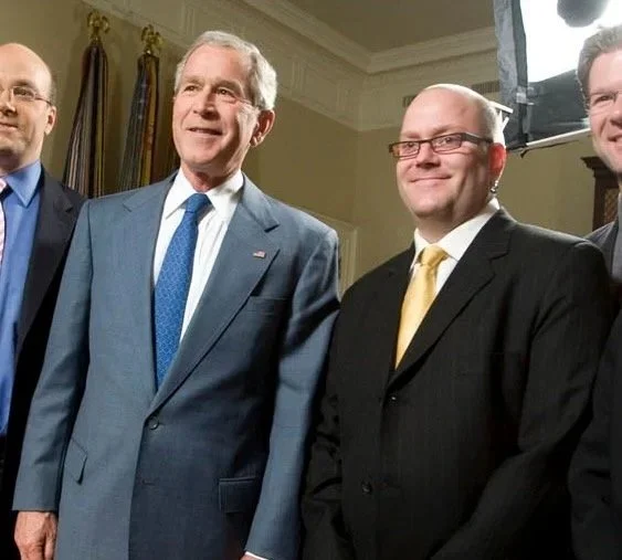 Group of four men in suits standing together indoors, with a man in the center wearing a blue suit and two men to his right in black suits and yellow ties, smiling and posing for a photo.