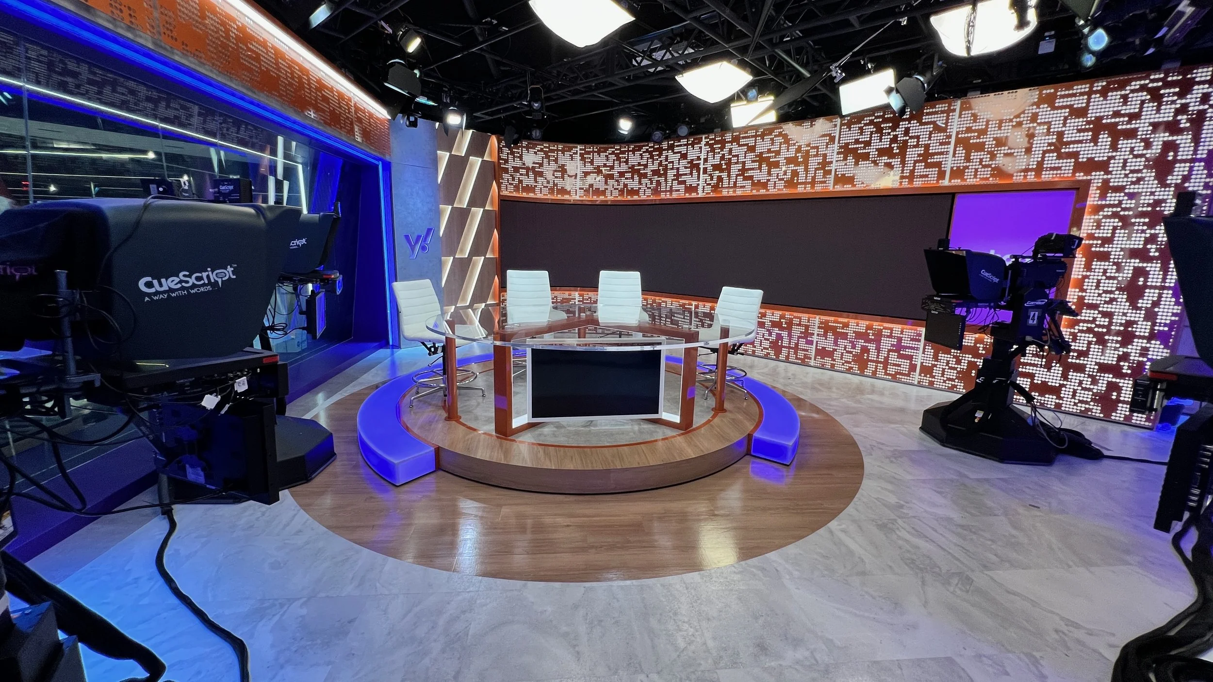 A television or news studio set with a round desk, four white chairs behind the desk, and multiple cameras and equipment around the studio. The background features large digital screens and ambient LED lighting, creating a modern, high-tech appearance.
