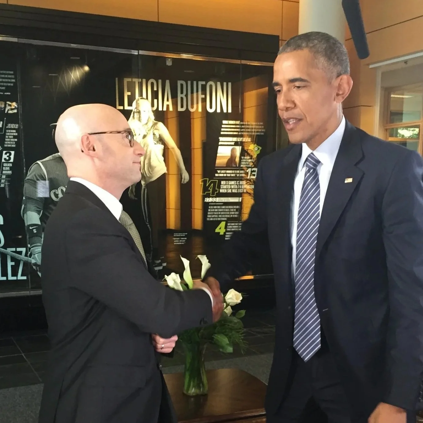 Two men in suits shaking hands, one holding white roses in a lobby with a display about Leticia Buffoni in the background.