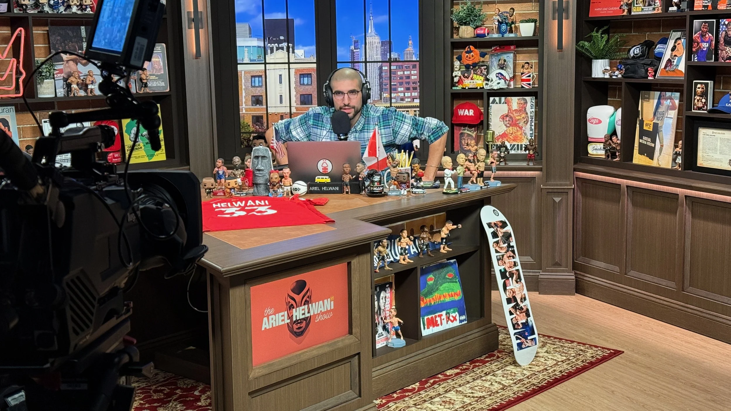 A man with a shaved head, glasses, and a plaid shirt is speaking into a microphone at a podcast studio filled with figurines, books, and decorations, with a city skyline visible through the window behind him.