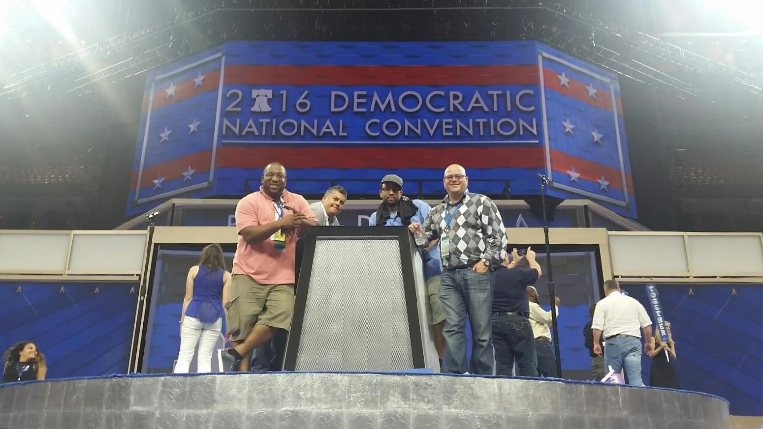 Four men standing on a stage in front of a large screen displaying '2016 Democratic National Convention', with other convention attendees visible in the background.