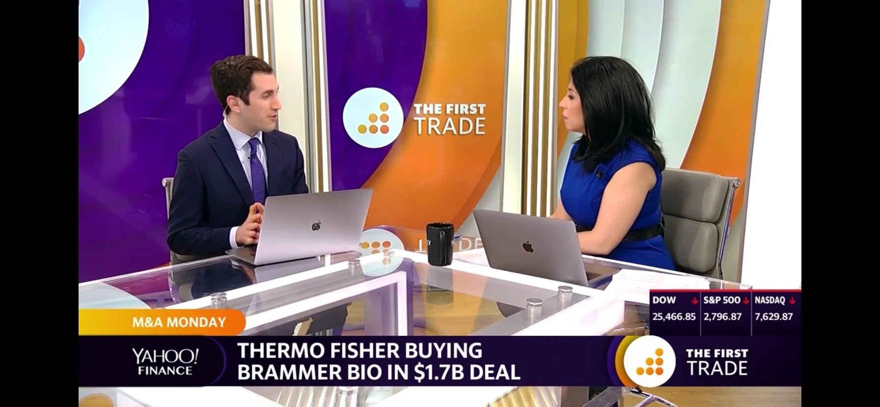 Two people, a man and a woman, are seated at a desk during a TV interview about Thermo Fisher buying Brammer Bio in a $1.7 billion deal. The man has short dark hair and is wearing a dark suit and purple tie. The woman has shoulder-length black hair and is wearing a blue dress. There are laptops, a mug, and papers on the desk. The background features a purple and orange set with the logo and branding for "The First Trade" and the Yahoo Finance logo.