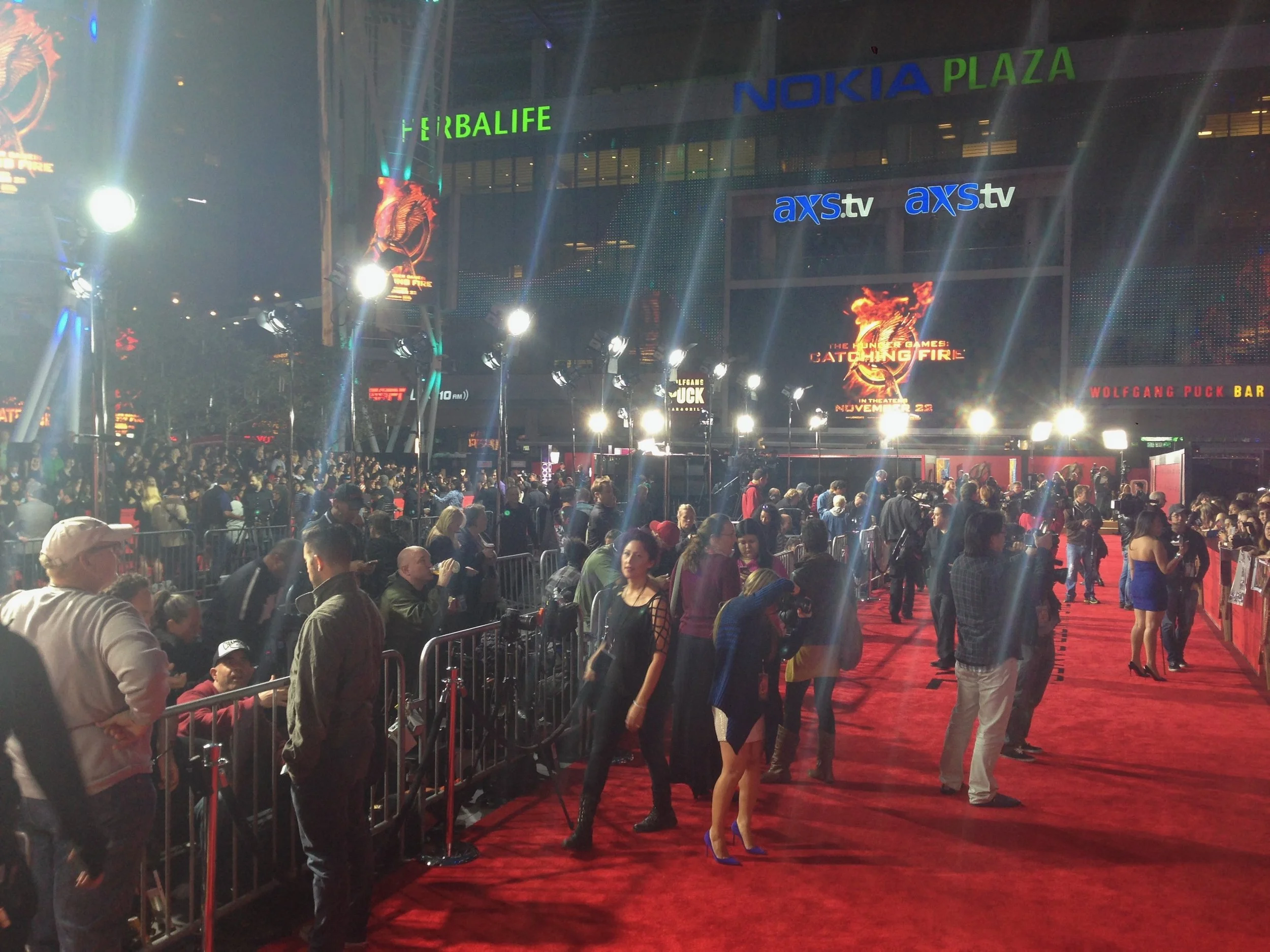 Crowd of people standing in line at an indoor event with large illuminated signs and banners for 'Nokia Plaza,' 'Herbalife,' and promotional displays for movies such as 'The Hunger Games: Catching Fire.' Red carpet and barriers are also visible.