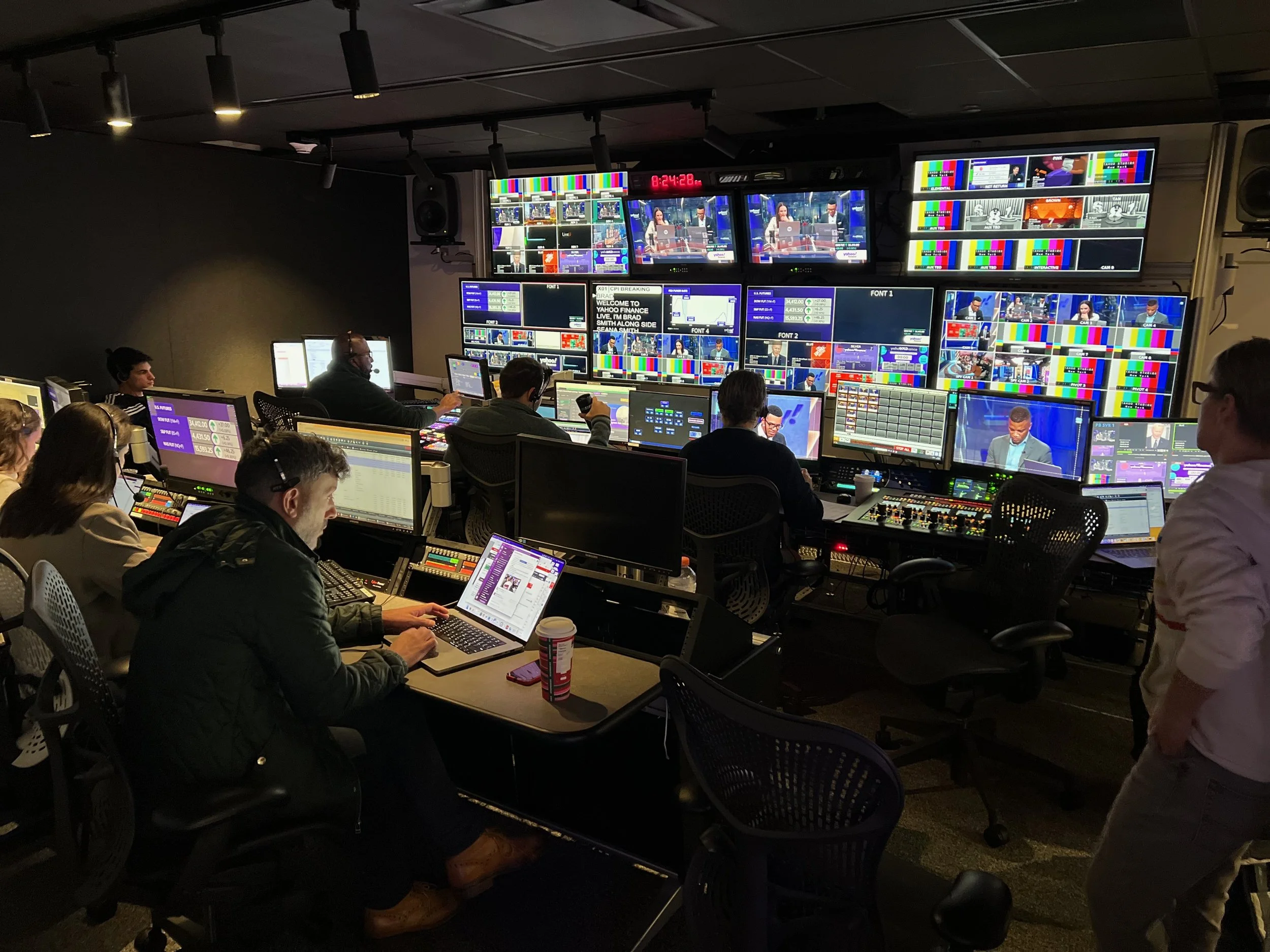 A control room with multiple monitors displaying live broadcast footage of a news program, with technicians working at their desks and people walking around.