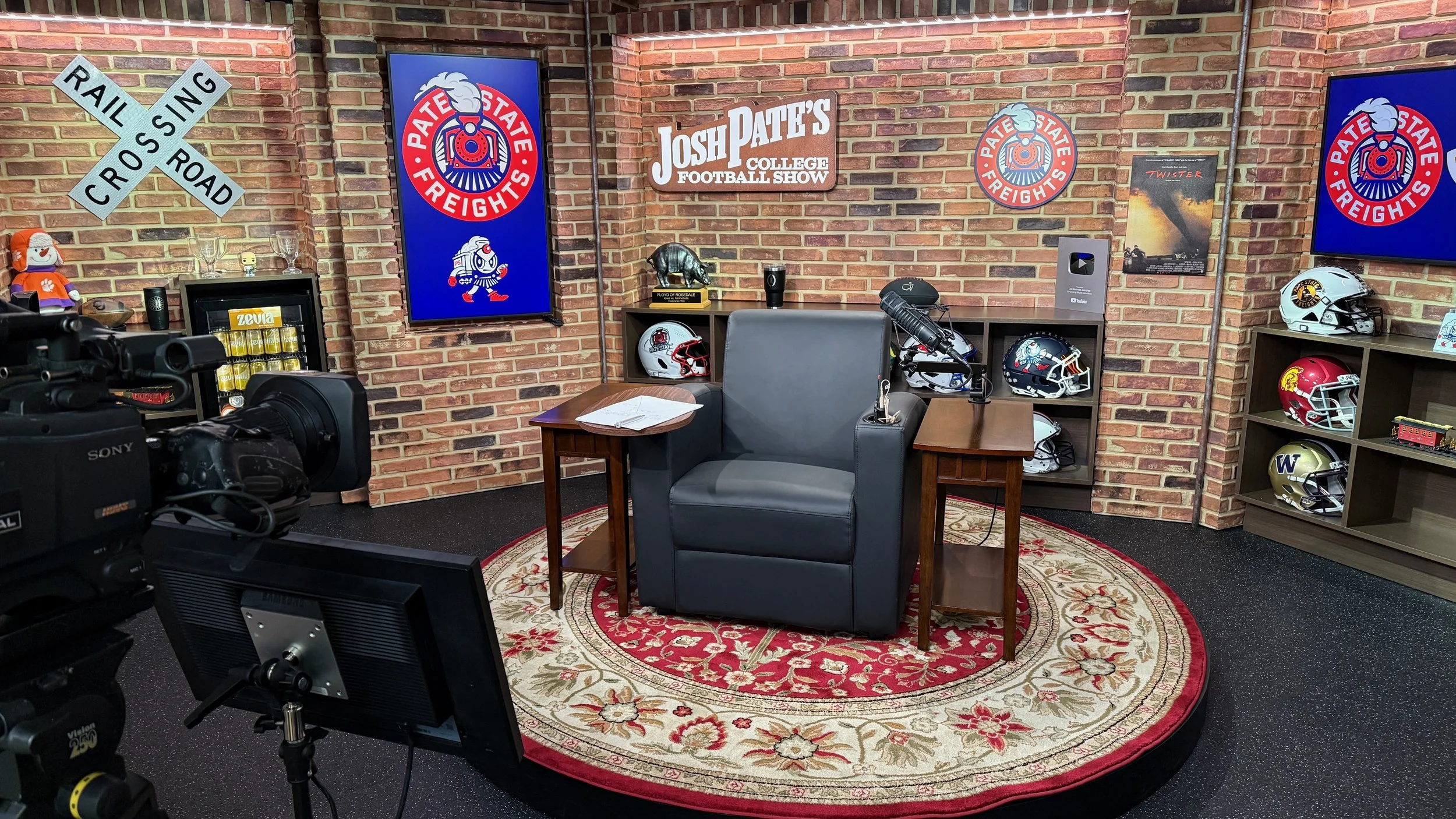 A television studio set decorated with brick walls and football helmets from various teams on shelves. There's a large rectangular sign reading 'Josh Pate's College Football Show' and two posters about 'Patriot State' and 'FREIGHTS.' A gray armchair and a small wooden table with papers are in the center, and a camera is positioned in front.