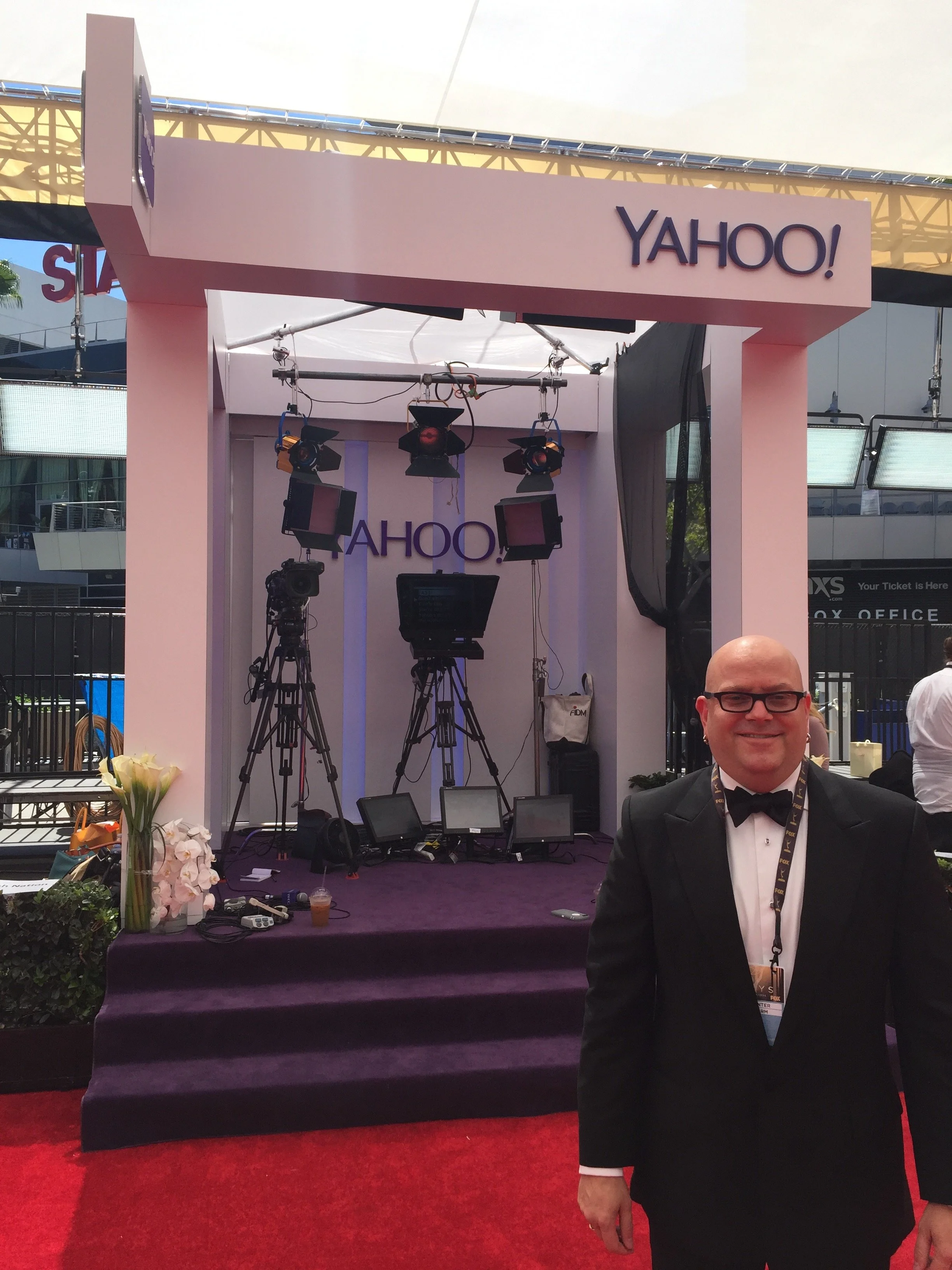A man in a tuxedo with glasses posing at a Yahoo event, standing on a red carpet in front of a stage with cameras and lighting equipment.