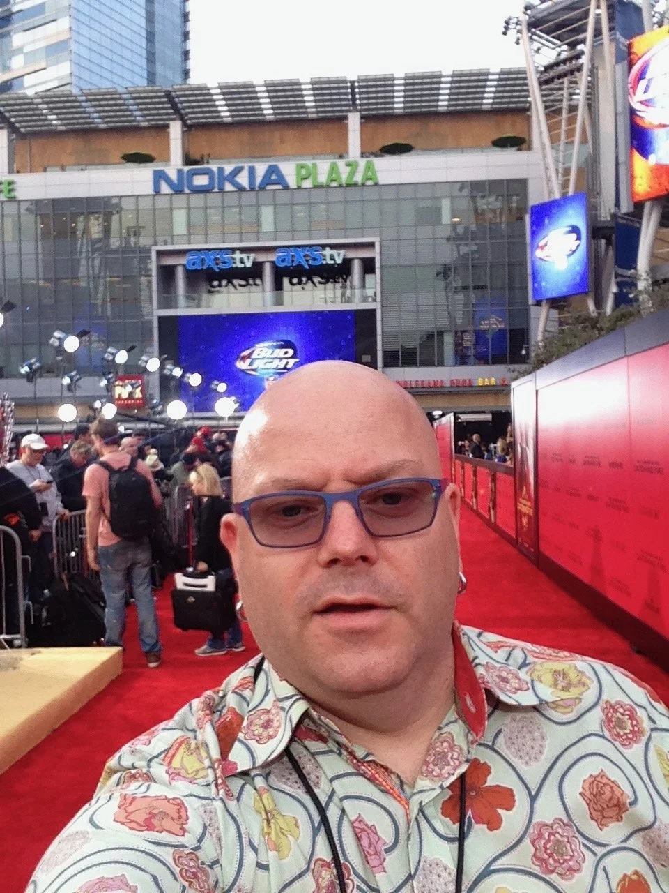 A man taking a selfie at an outdoor event with a large crowd in the background. The backdrop features a branded building with signs for Nokia Plaza, axs.tv, and Bud Light