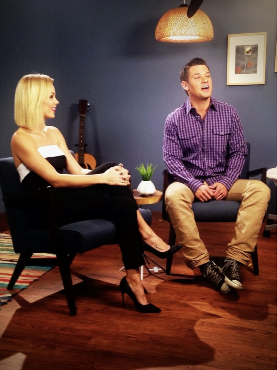 A woman and a man sitting in chairs in a room with a dark blue wall, a guitar in the background, and framed pictures. The woman is dressed in black heels and a strapless black and white outfit, smiling and looking at the man. The man is wearing a purple checkered shirt, beige pants, and sneakers, speaking or singing, with a lamp hanging above.