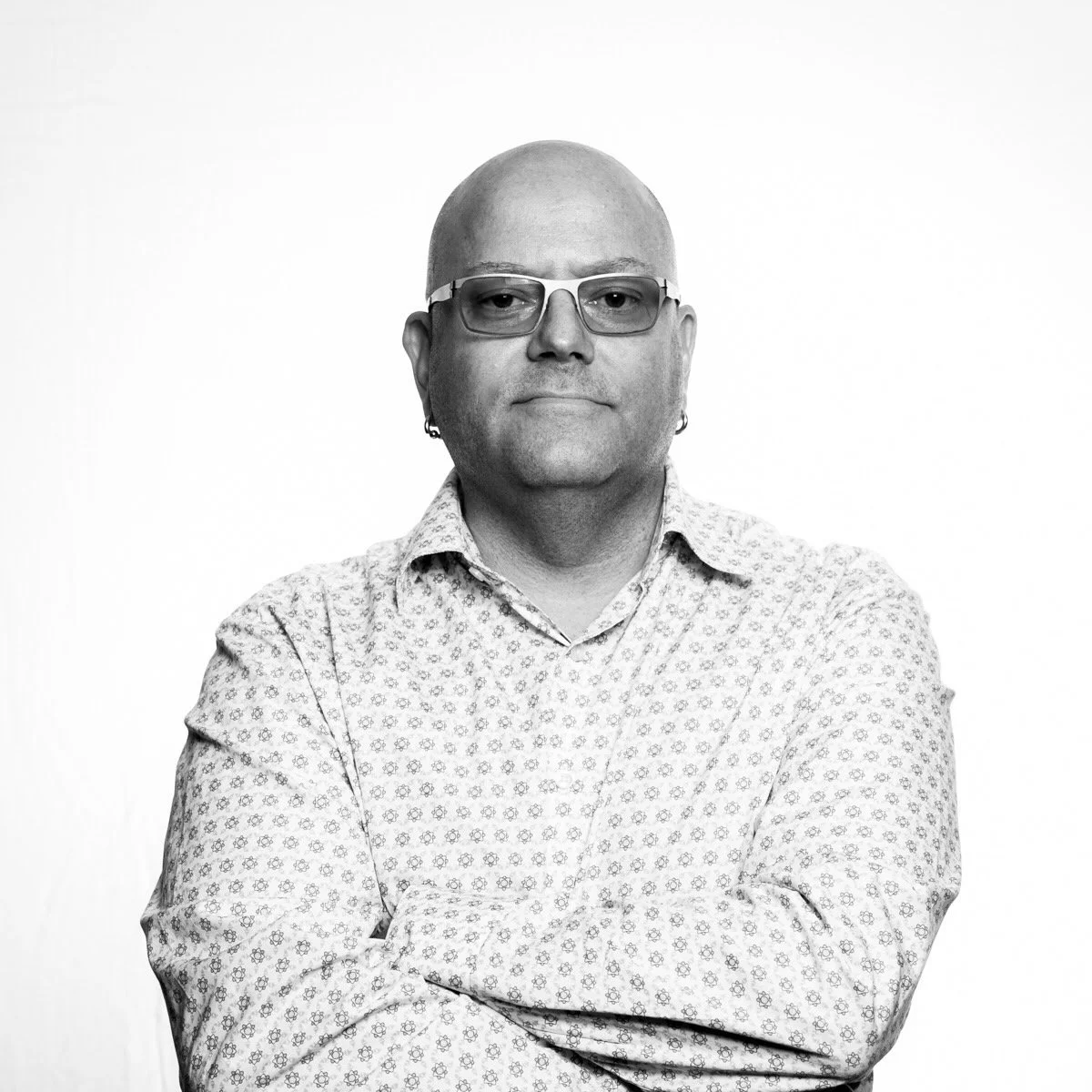 Black and white portrait of a middle-aged man with glasses, earrings, and a patterned shirt, standing with arms crossed against a plain background.