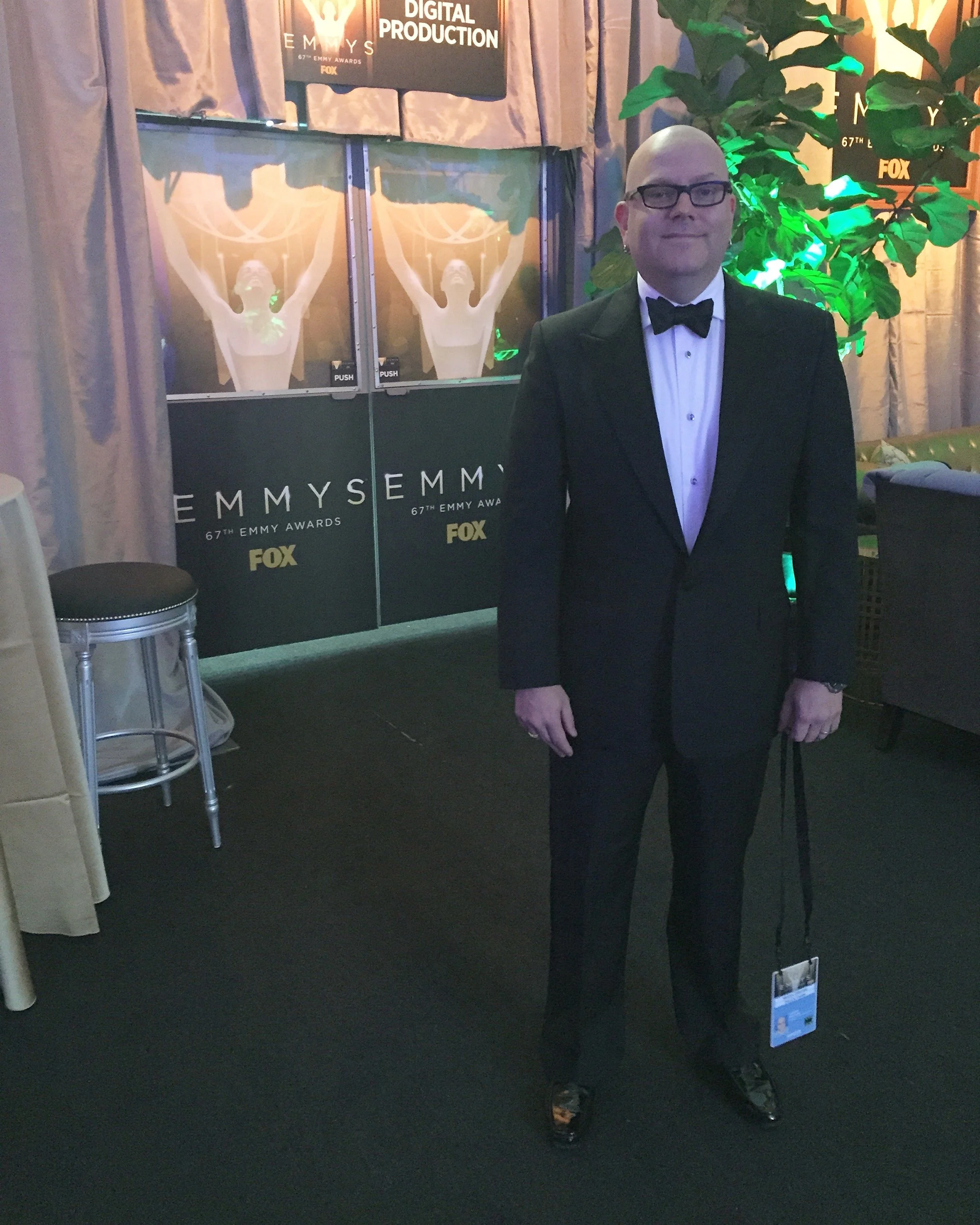 Man in tuxedo with glasses and a bow tie standing in front of Emmy Awards signage, with a large green plant on the right.