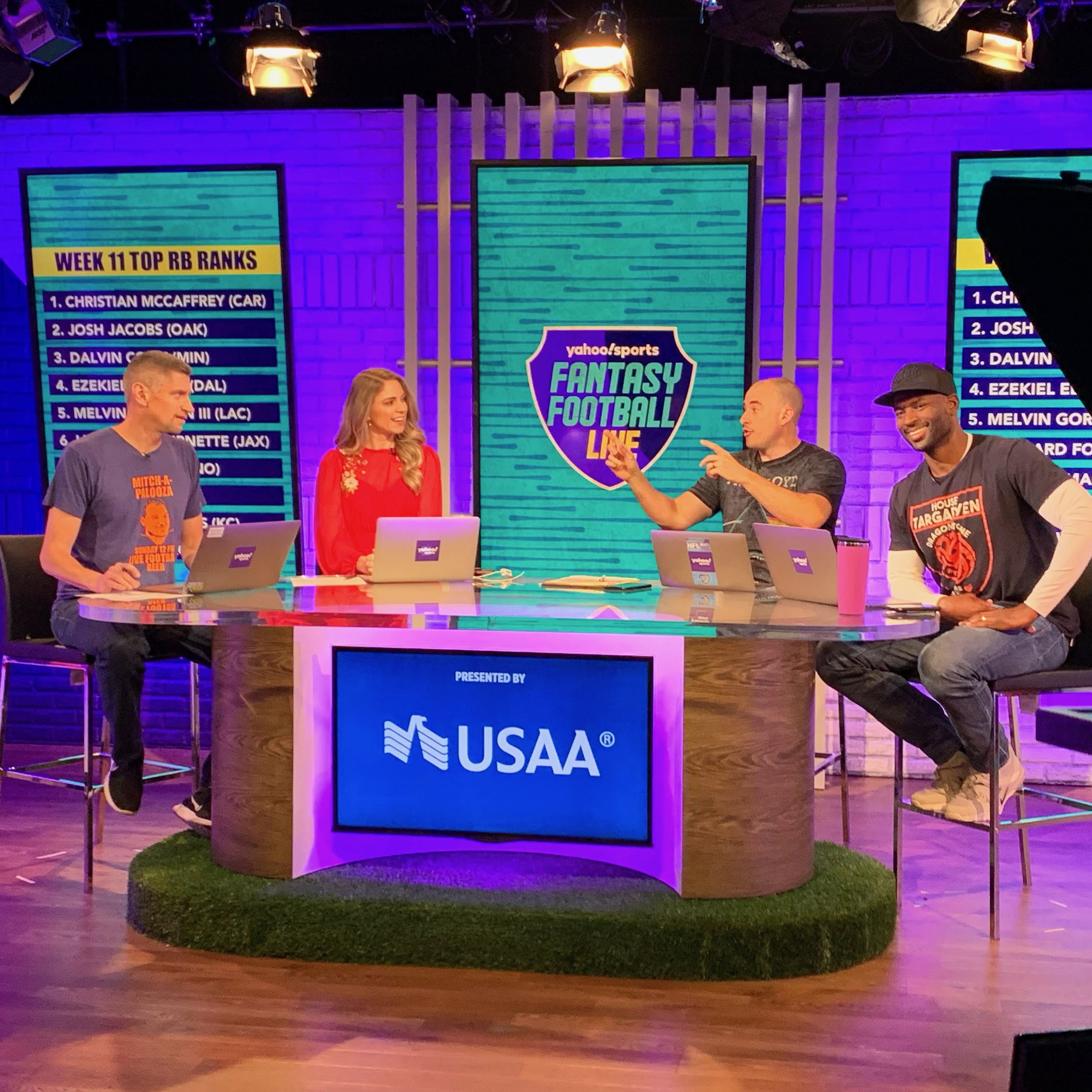 Indoor stage setup with large screens displaying 'Week 11 Top RB Ranks' and 'Yahoo Sports Fantasy Football Line.' Four people are sitting and standing at the table, engaged in discussion, with a prominent USA Today logo on the front of the table. The background is lit with purple lighting.