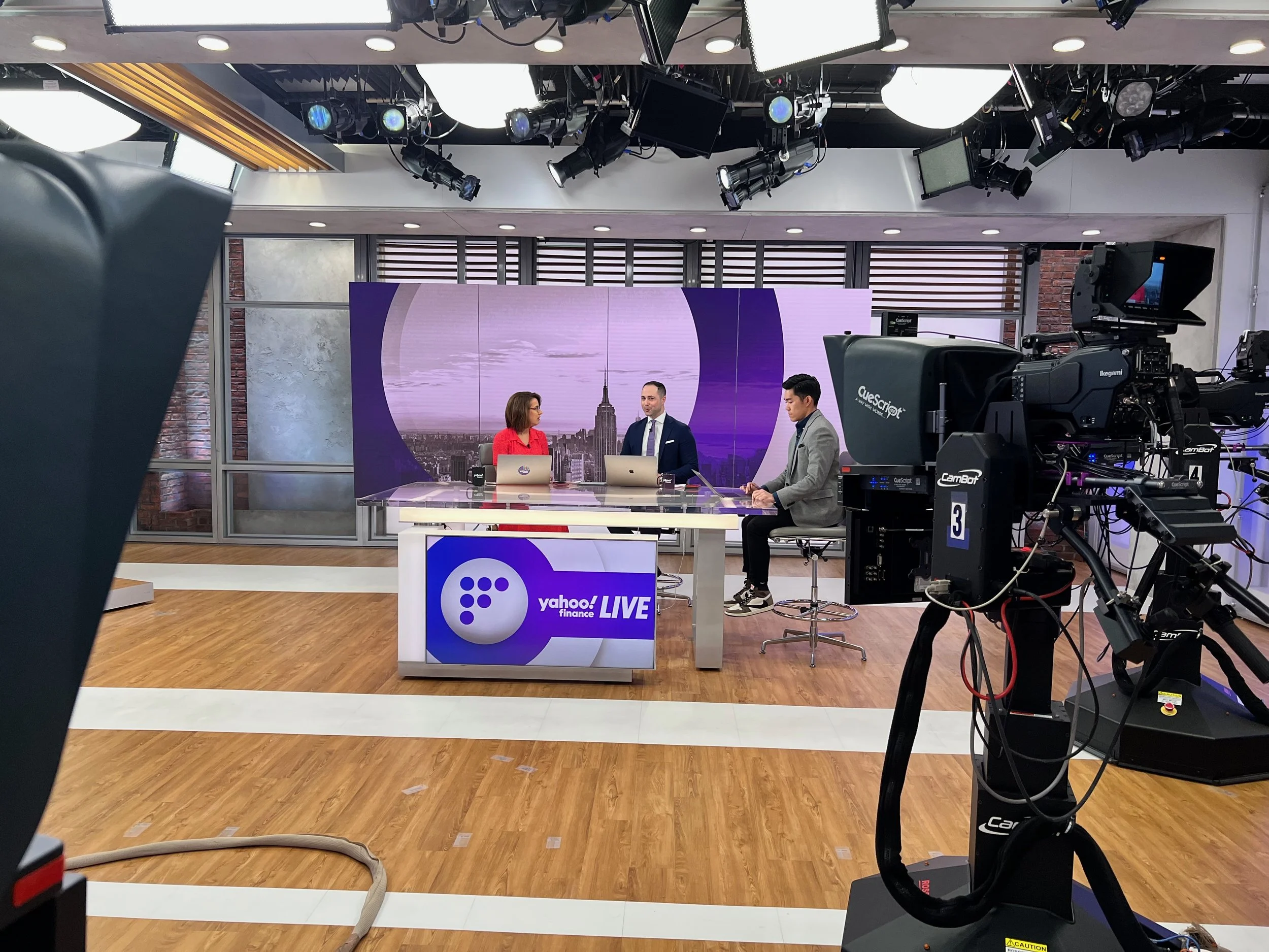 News studio with three anchors seated at a desk, in front of a large screen displaying the New York City skyline with the Empire State Building, surrounded by studio lighting, cameras, and production equipment.