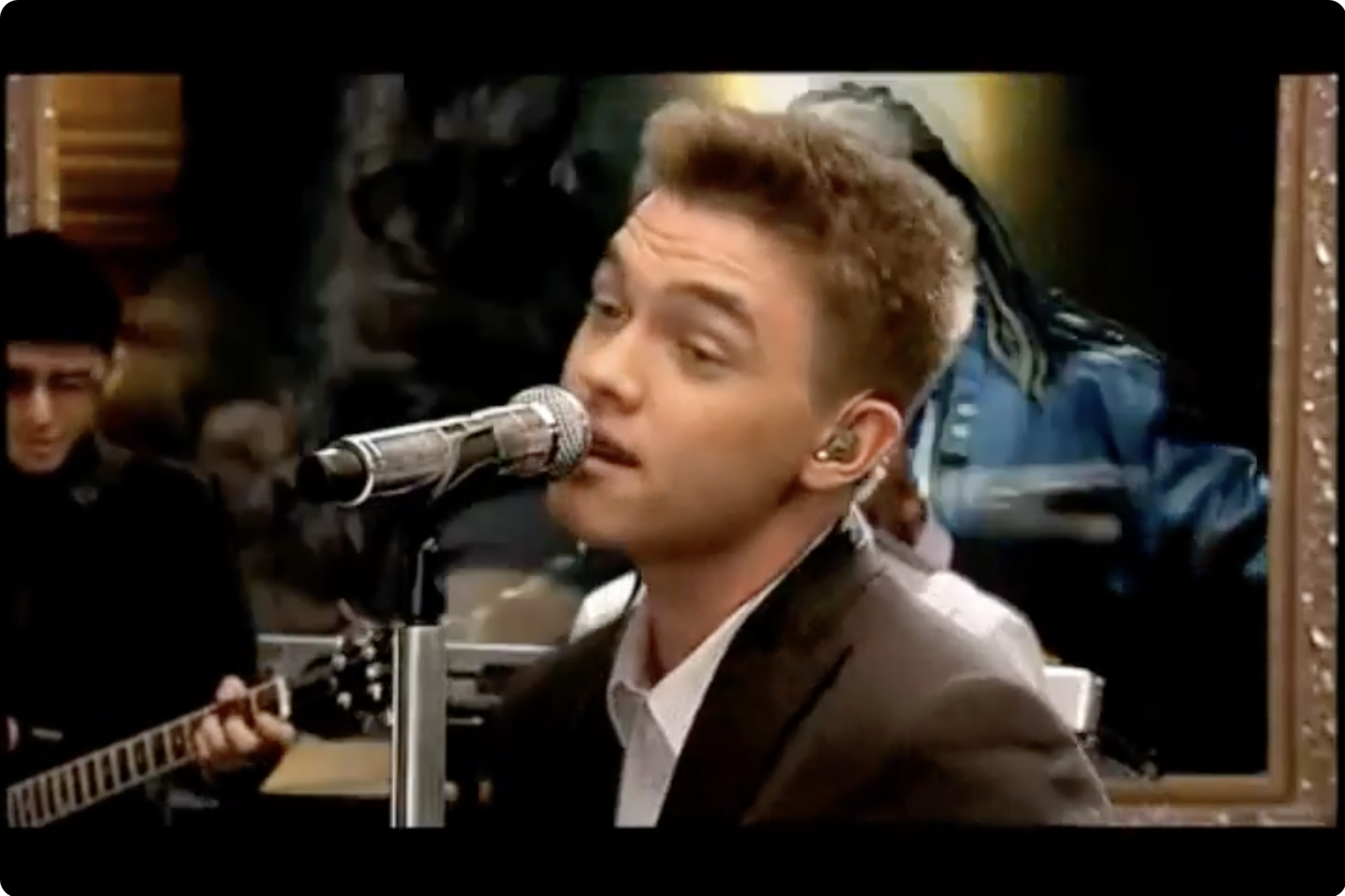 A young man singing into a microphone while playing an acoustic guitar, with a second person playing guitar on the left in a lively indoor setting.