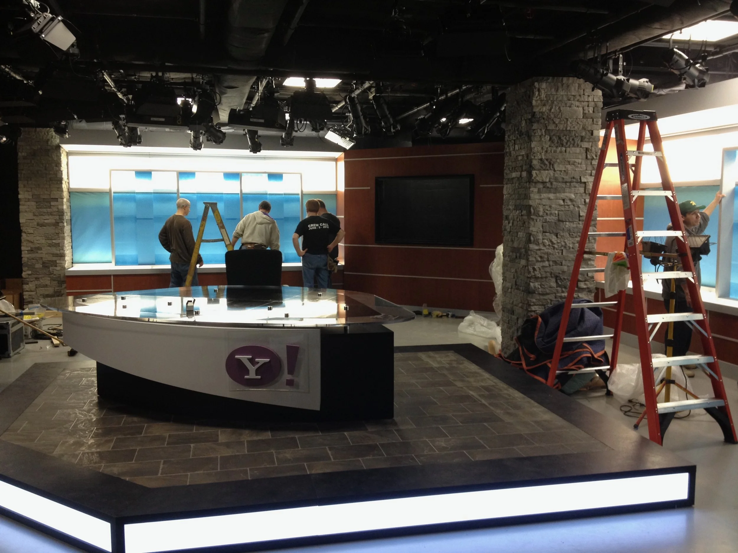 A television studio with crew members setting up lighting and equipment, a news desk with a purple 'Y!' logo, and a backdrop of blue and orange panels.
