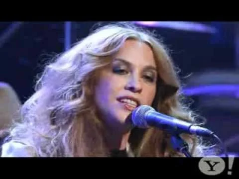 A woman with long curly blonde hair singing into a microphone on stage.
