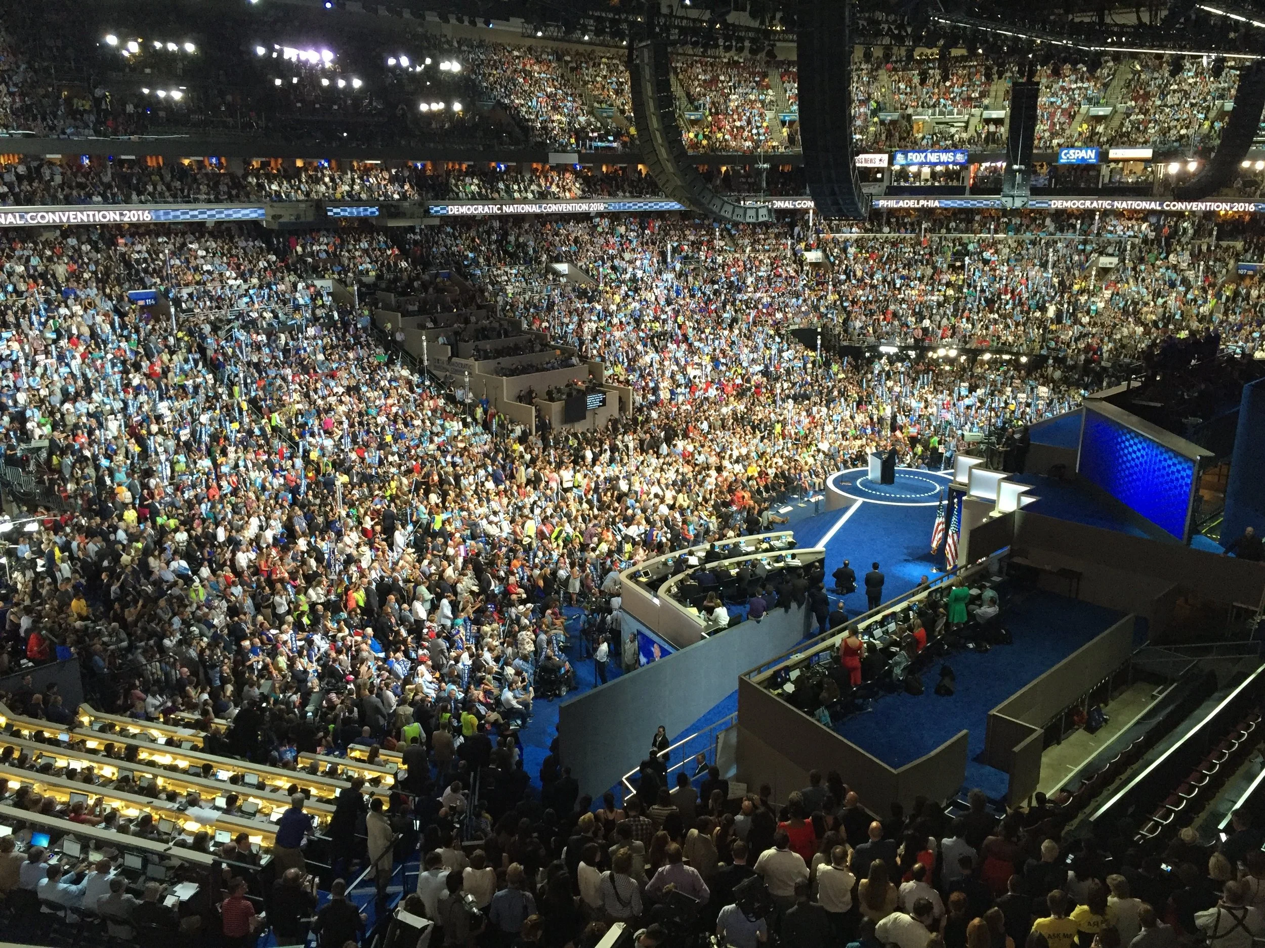 Crowd of people attending the Democratic National Convention 2016 in Philadelphia, inside a large stadium with multiple levels of seating, stage, and screens displaying event information.