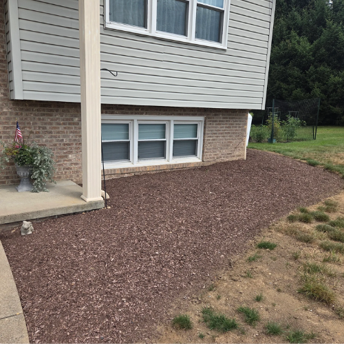 A small area of bare soil with mulch around the exterior of a house, adjacent to a concrete sidewalk and a grassy lawn, with a potted plant and an American flag nearby.