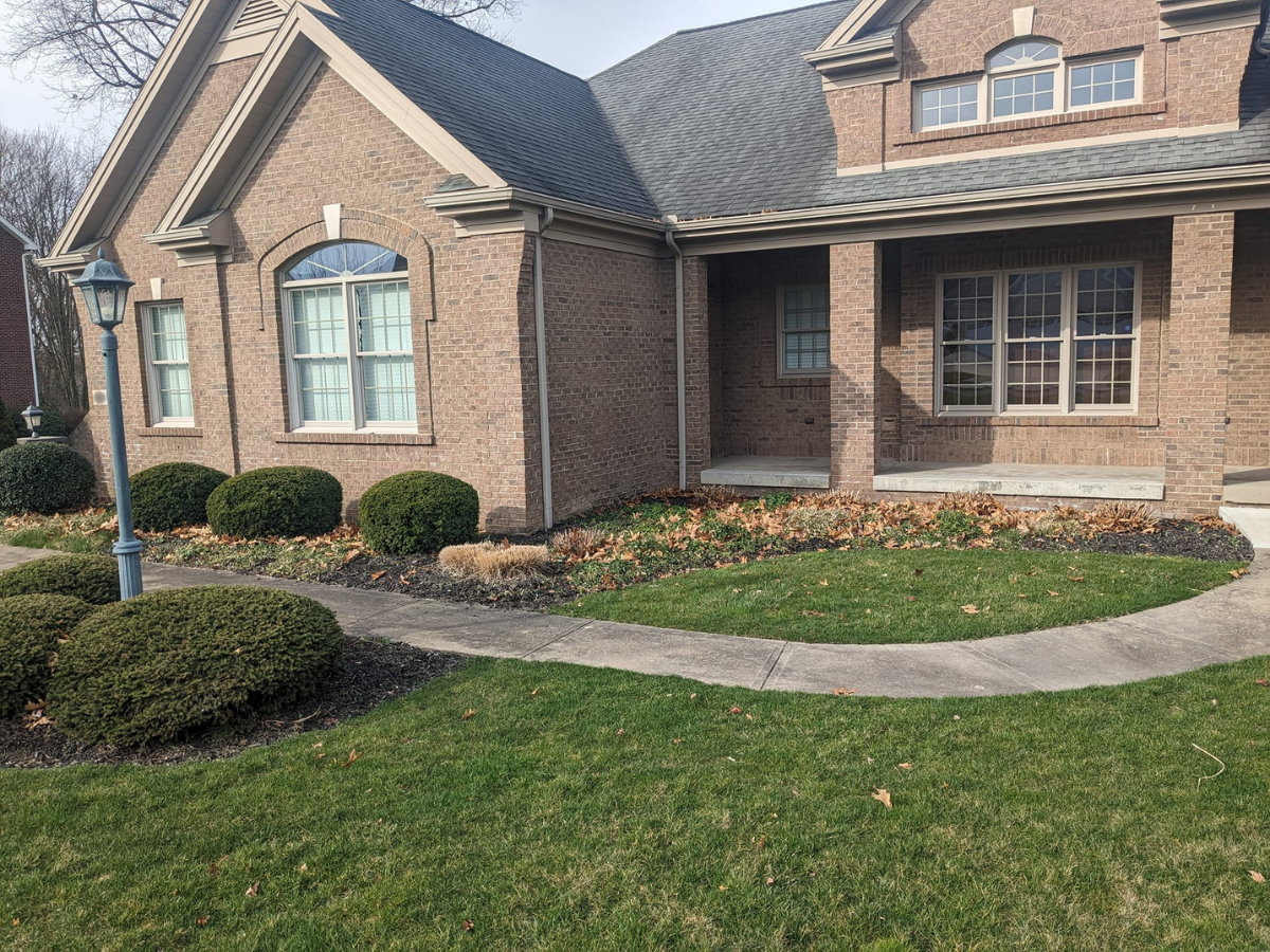 Front yard of a brick house with a curved concrete walkway, green lawn, bushes, and a lamp post.